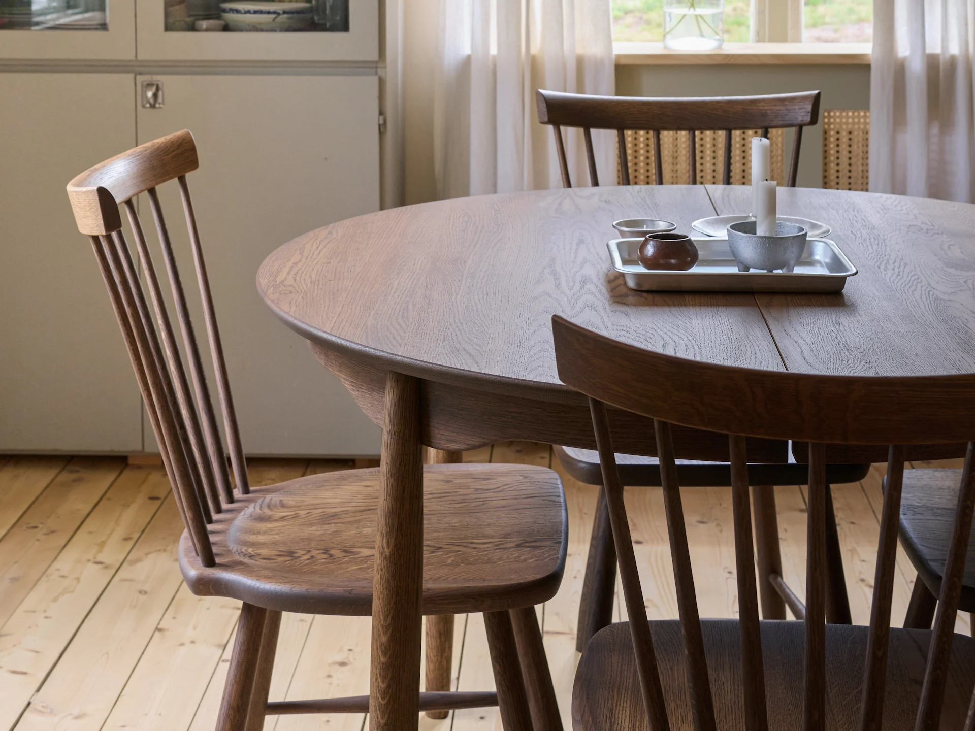 Een ronde houten eettafel met drie stoelen met spijlenrug, een dienblad met kaarsen en een kom, en een houten vloer.
