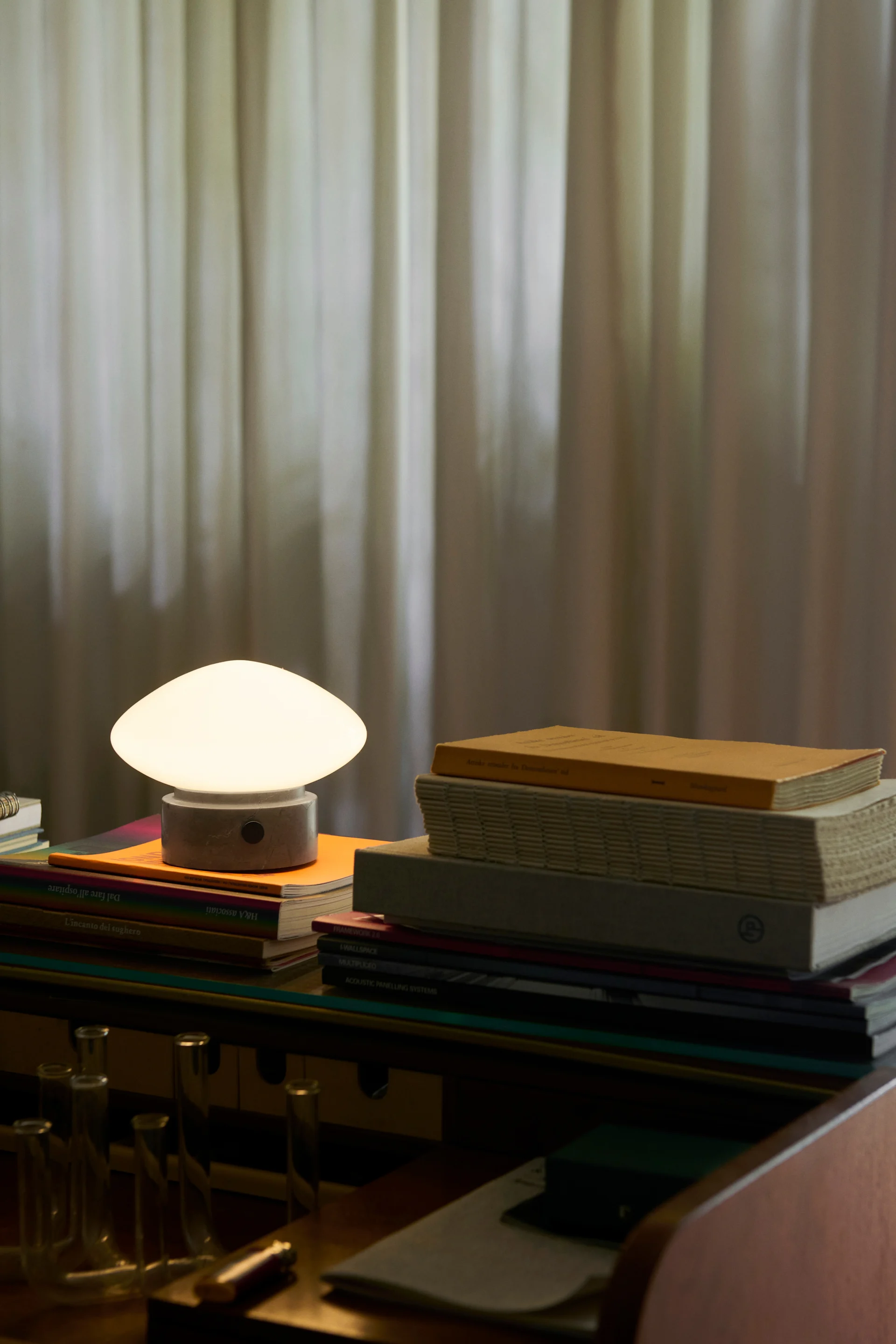 Paddenstoelvormige tafellamp met een gloeiende witte koepel en grijze voet, staand op boeken op een houten bureau.