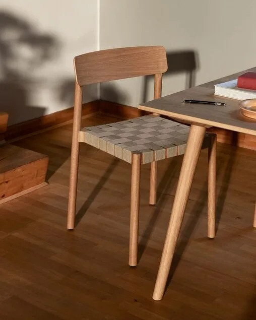 Een lichte houten stoel met een geweven geruite zitting naast een tafel met boeken en een pen.