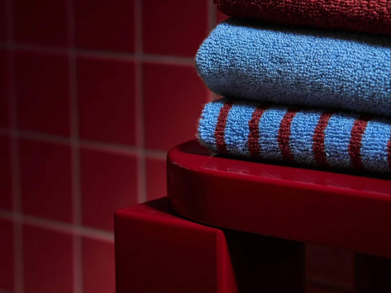 Stapel van drie gevouwen handdoeken: donkerrood, lichtblauw en blauw met rode strepen, op een rood oppervlak.