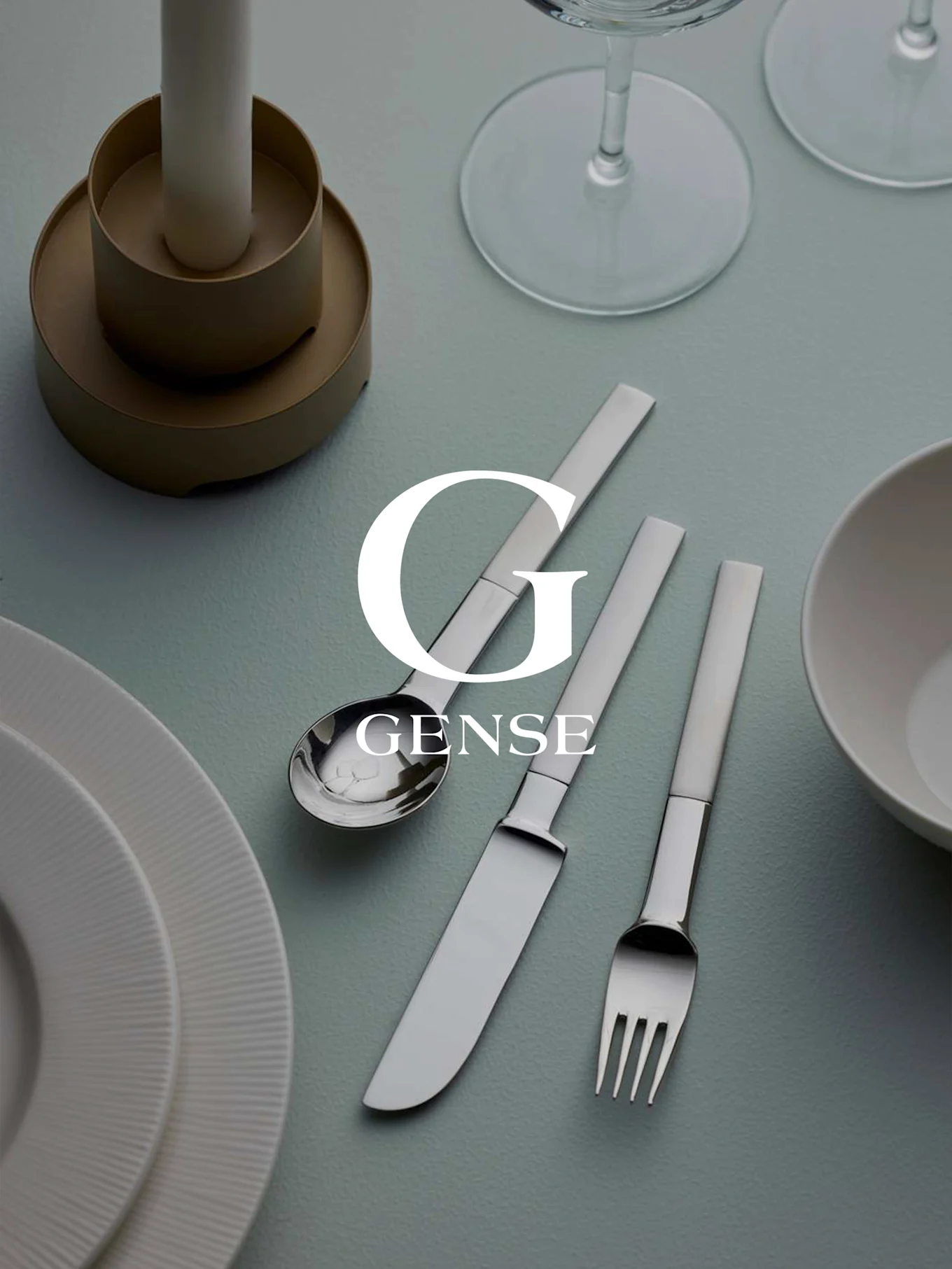 Een tafelschikking met een Gense zilveren bestekset (lepel, mes, vork), witte borden, een kaars en glazen op een lichtblauwe ondergrond.