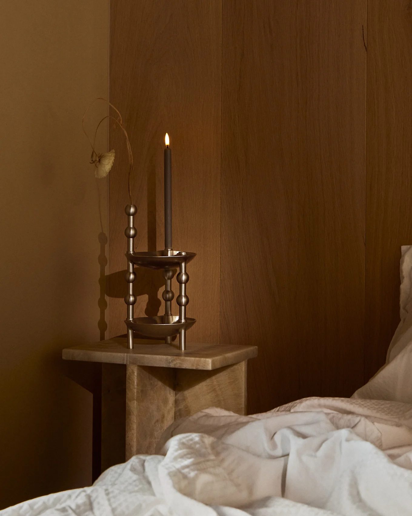 Een brandende donkere kaars in een sierlijke metalen houder op een houten nachtkastje naast een bed met wit beddengoed in een schemerige kamer.