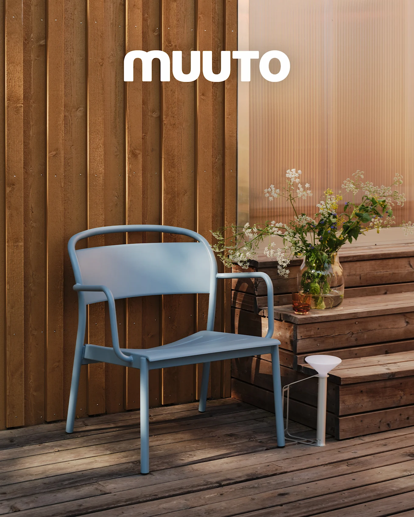 Een lichtblauwe buitenstoel op een houten terras naast treden met een vaas wilde bloemen en een witte draagbare lamp. MUUTO-logo.