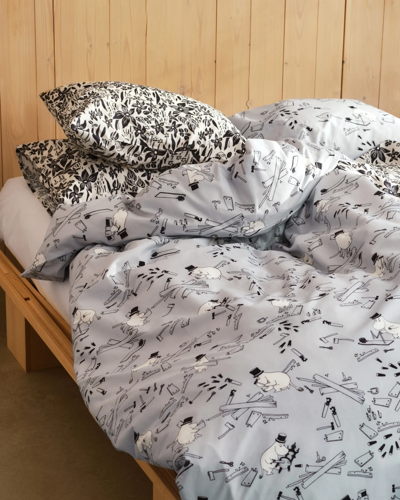 Bed met kussens in zwart-witte bloemenprint en een lichtblauw dekbed met cartoonfiguren, gereedschap en houten planken. Houten bedframe.