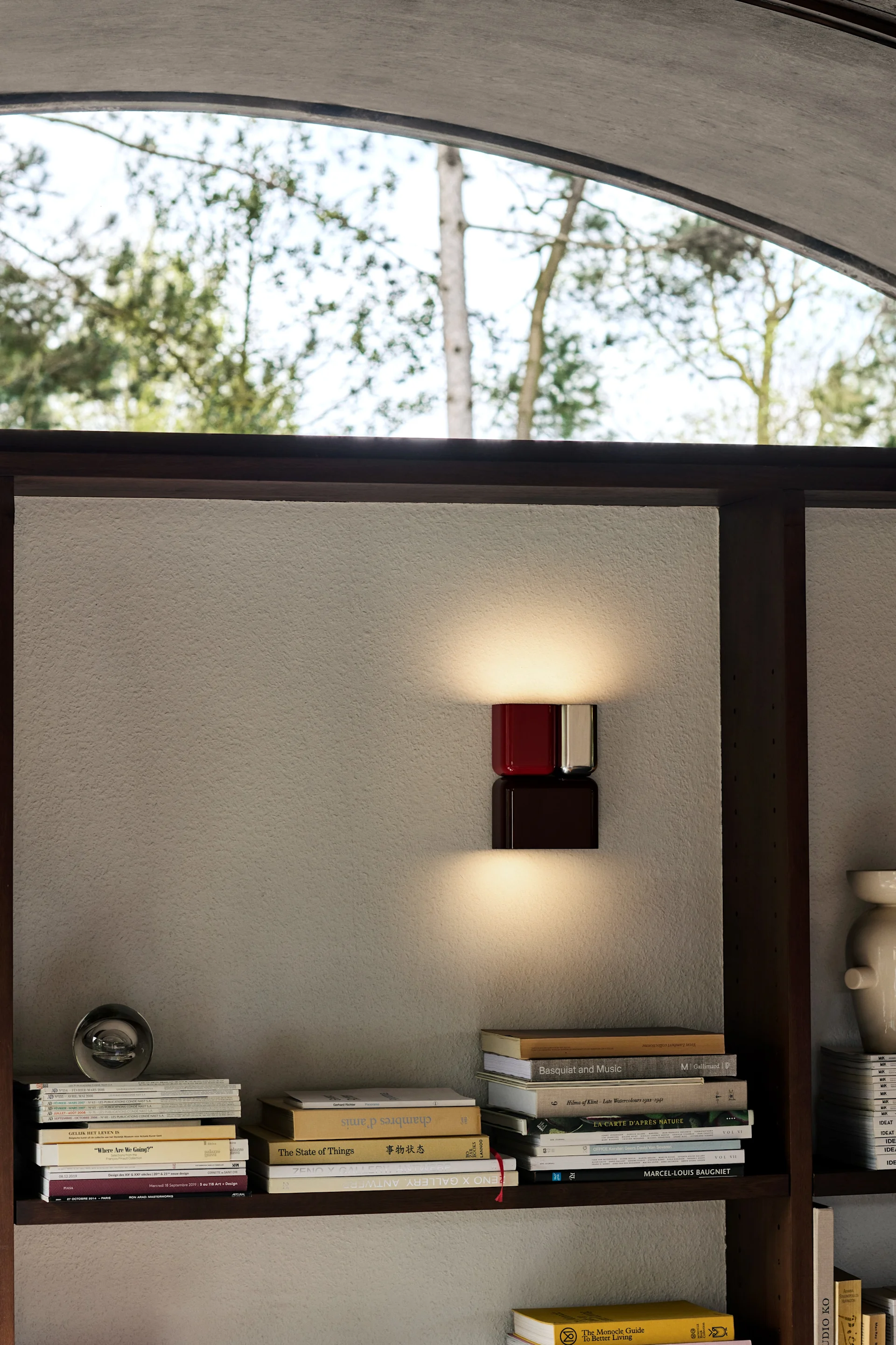 Een moderne wandlamp met een rood bovenste en een donkerbruin onderste kubusachtig element verlicht een gestructureerde muur tussen boekenplanken vol boeken.