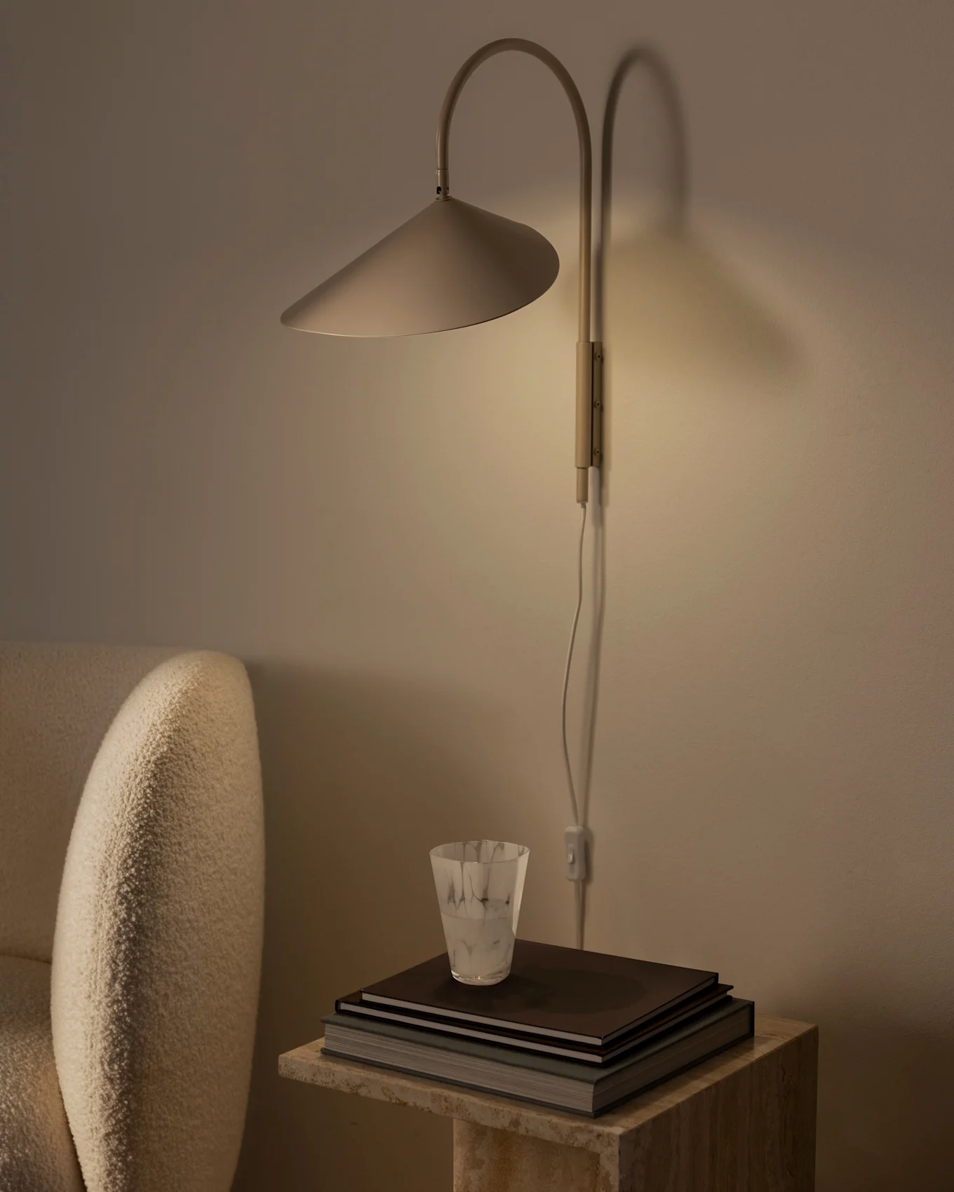 Warm verlichte wandlamp met conische kap boven een stapel boeken en een gestructureerd glas op een stenen bijzettafel, naast een beige fauteuil.