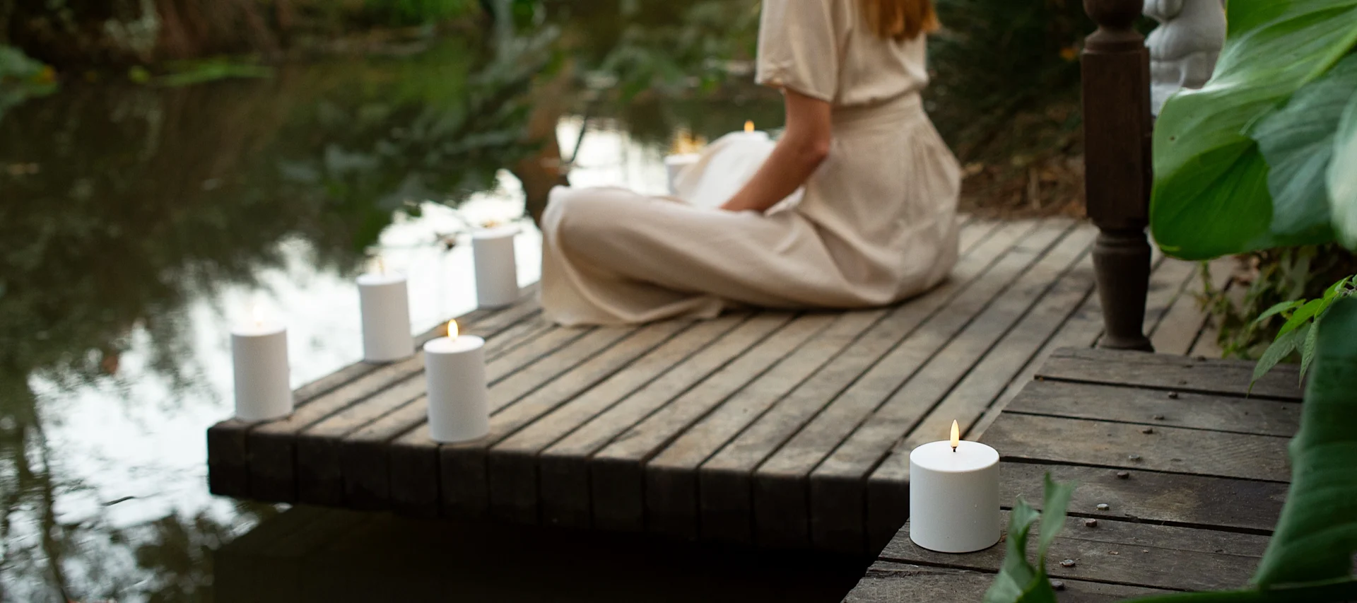 Een persoon in lichte kleding zit op een houten vlonder aan het water, omringd door meerdere brandende witte pilaarkaarsen.