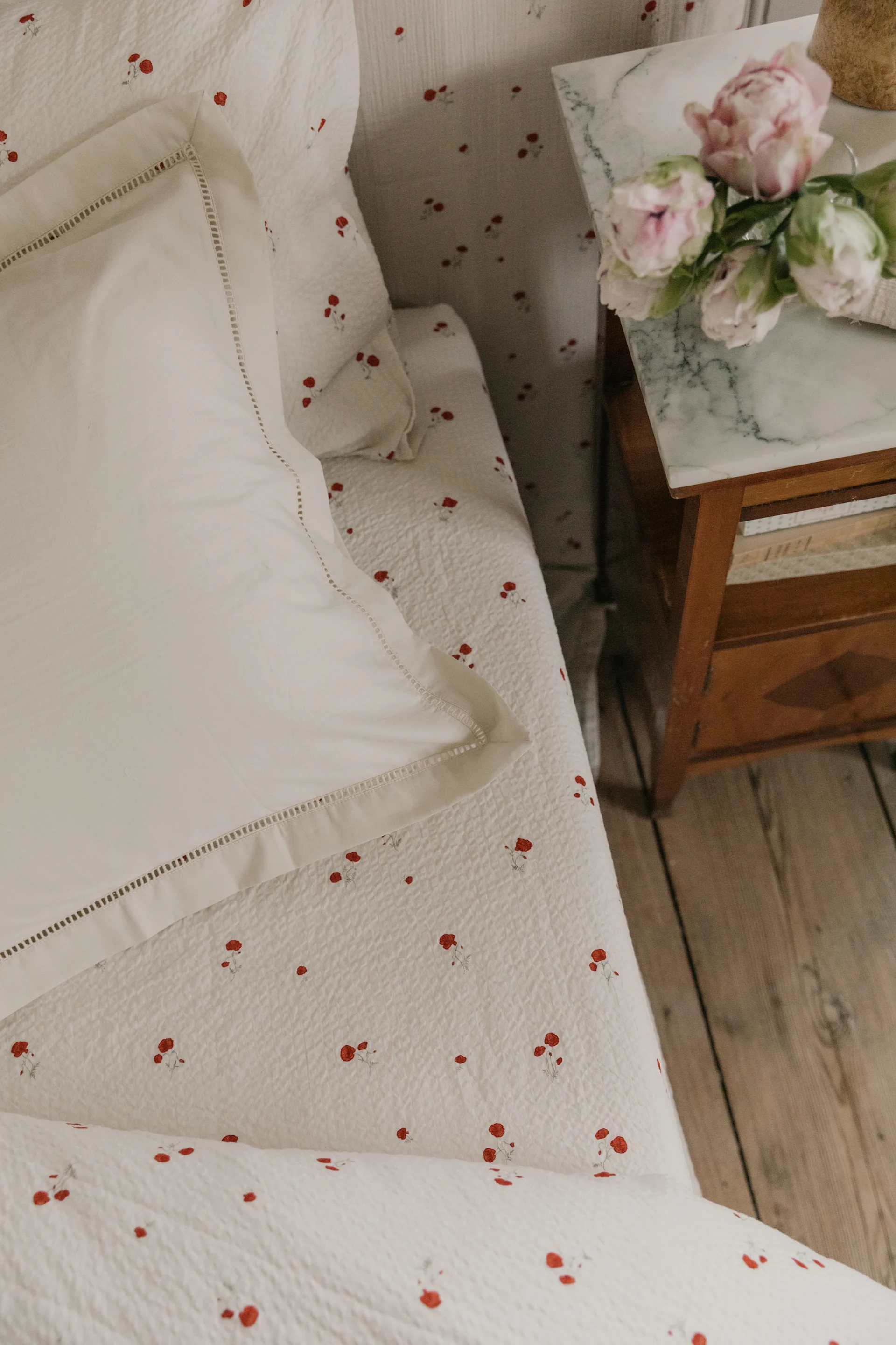 Wit beddengoed met rood bloemenpatroon, een wit kussen en roze bloemen op een marmeren nachtkastje.