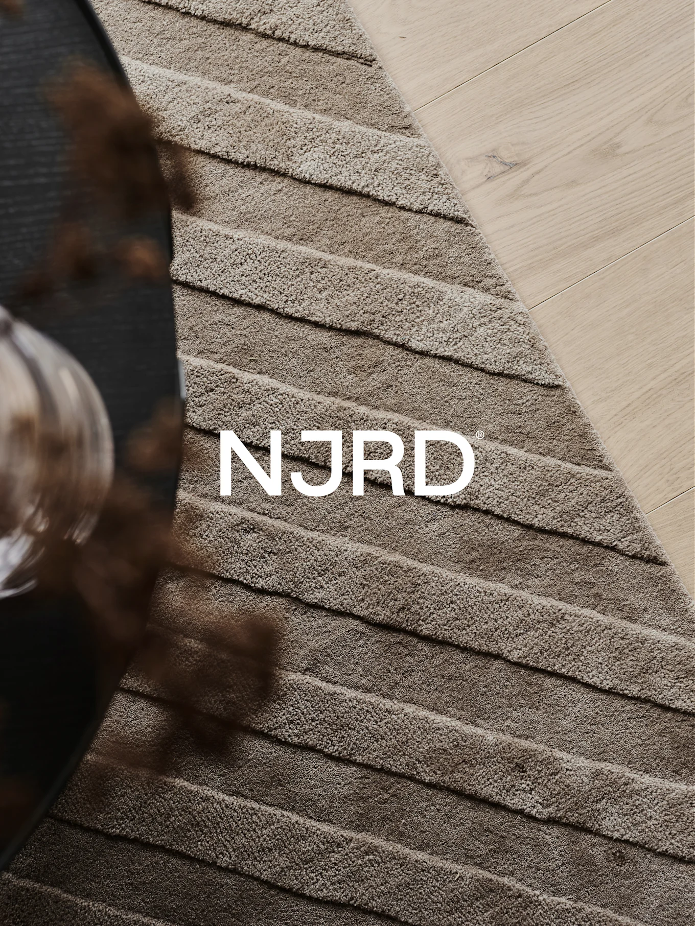 Close-up van een lichtbruin hoogpolig tapijt met horizontale ribbels, gedeeltelijk op een lichte houten vloer, met het witte 'NJRD'-logo.