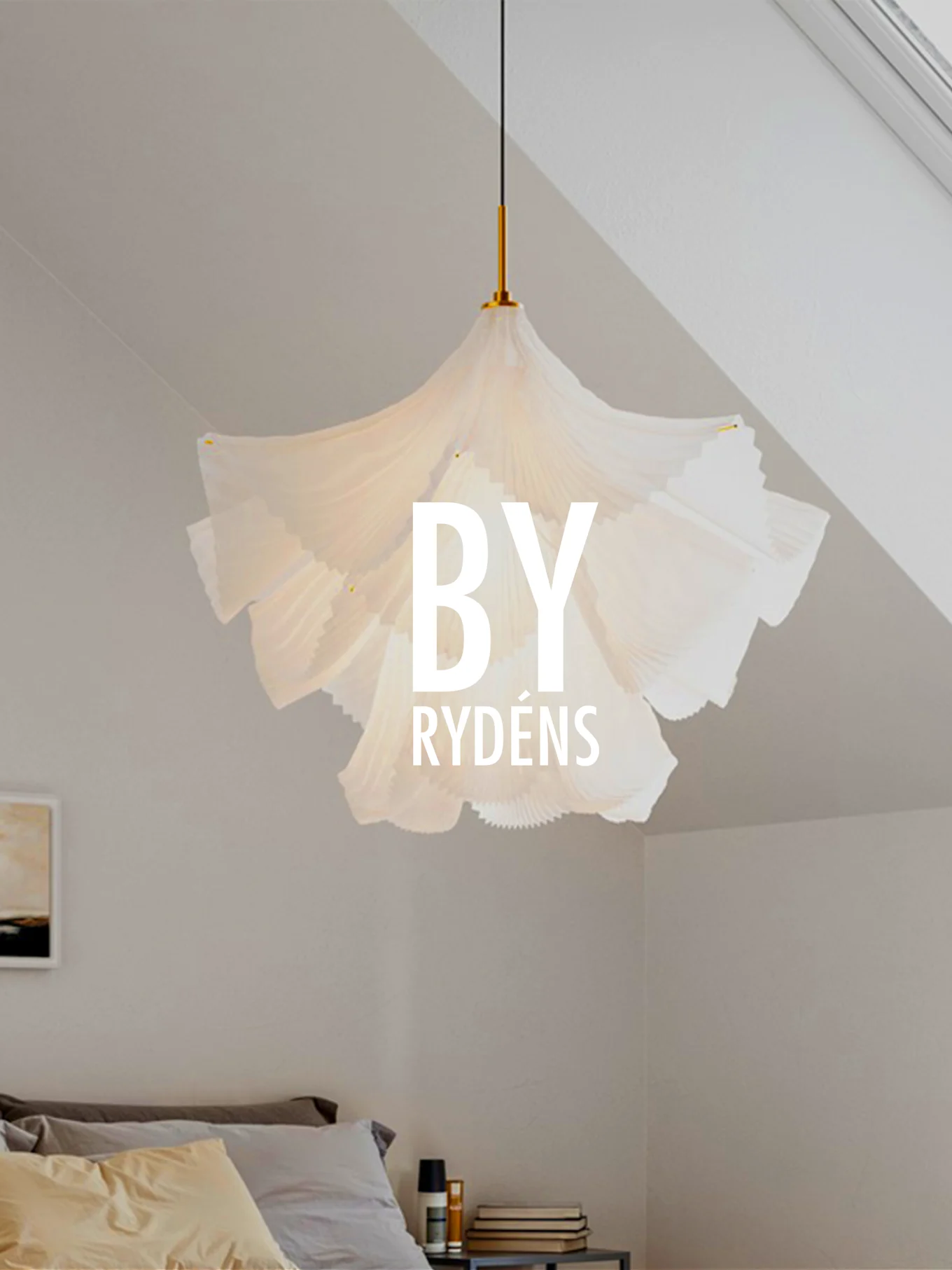 Een witte, geplooide sculpturale hanglamp hangt boven een bed met kussens en een nachtkastje. 'BY RYDÉNS' is over de lamp heen geplaatst.