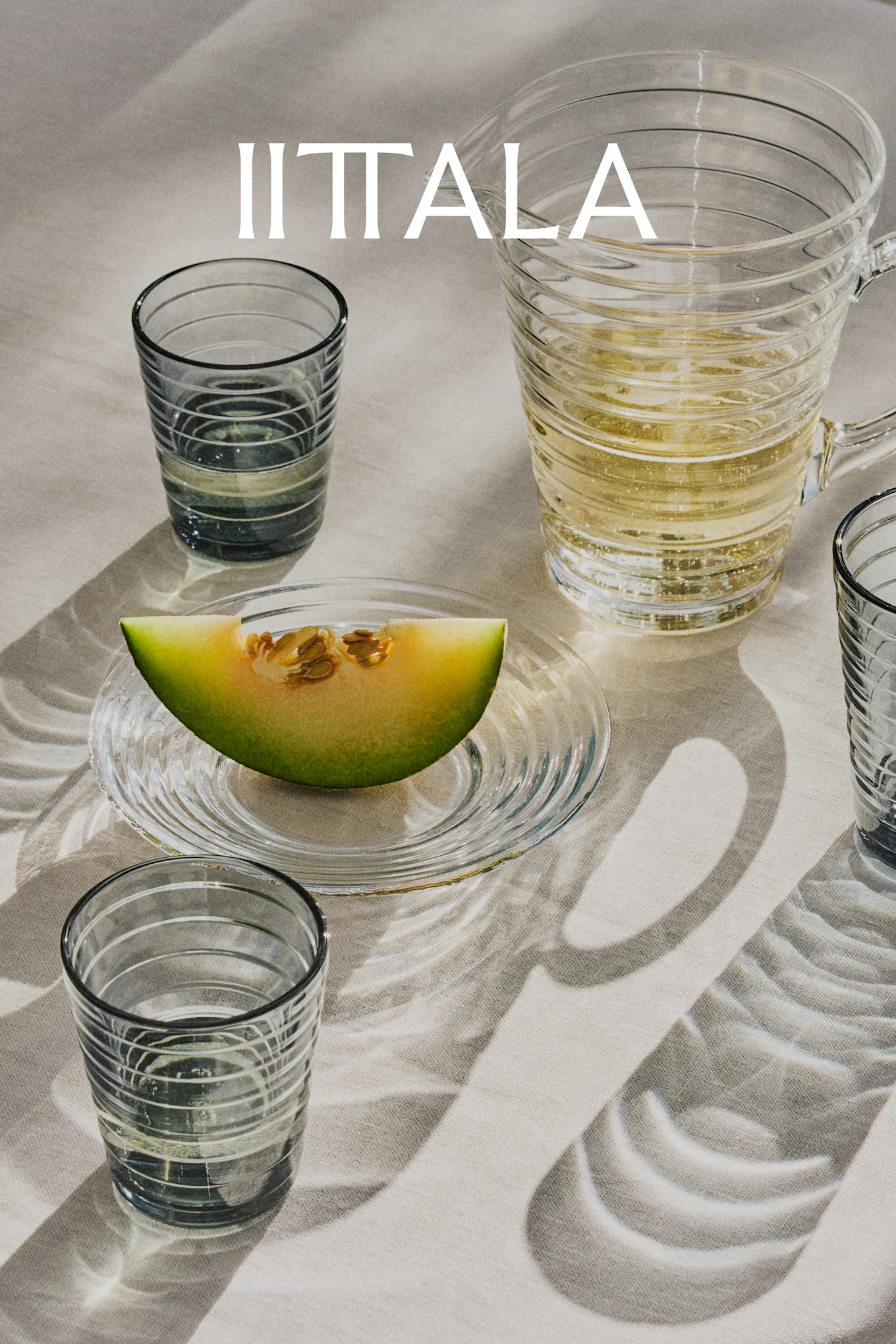 IITTALA-logo boven glazen, een kan met bruisende drank en een meloenschijf op een bord, alles op licht textiel.