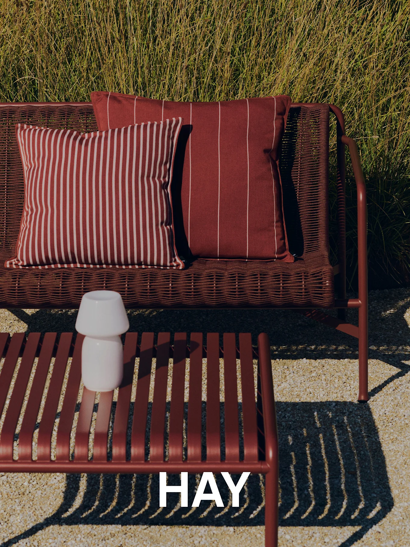 Buitenscène met een gevlochten rode bank, gestreepte kussens, een salontafel met latten en een witte lamp, en hoog gras op de achtergrond. HAY-logo.