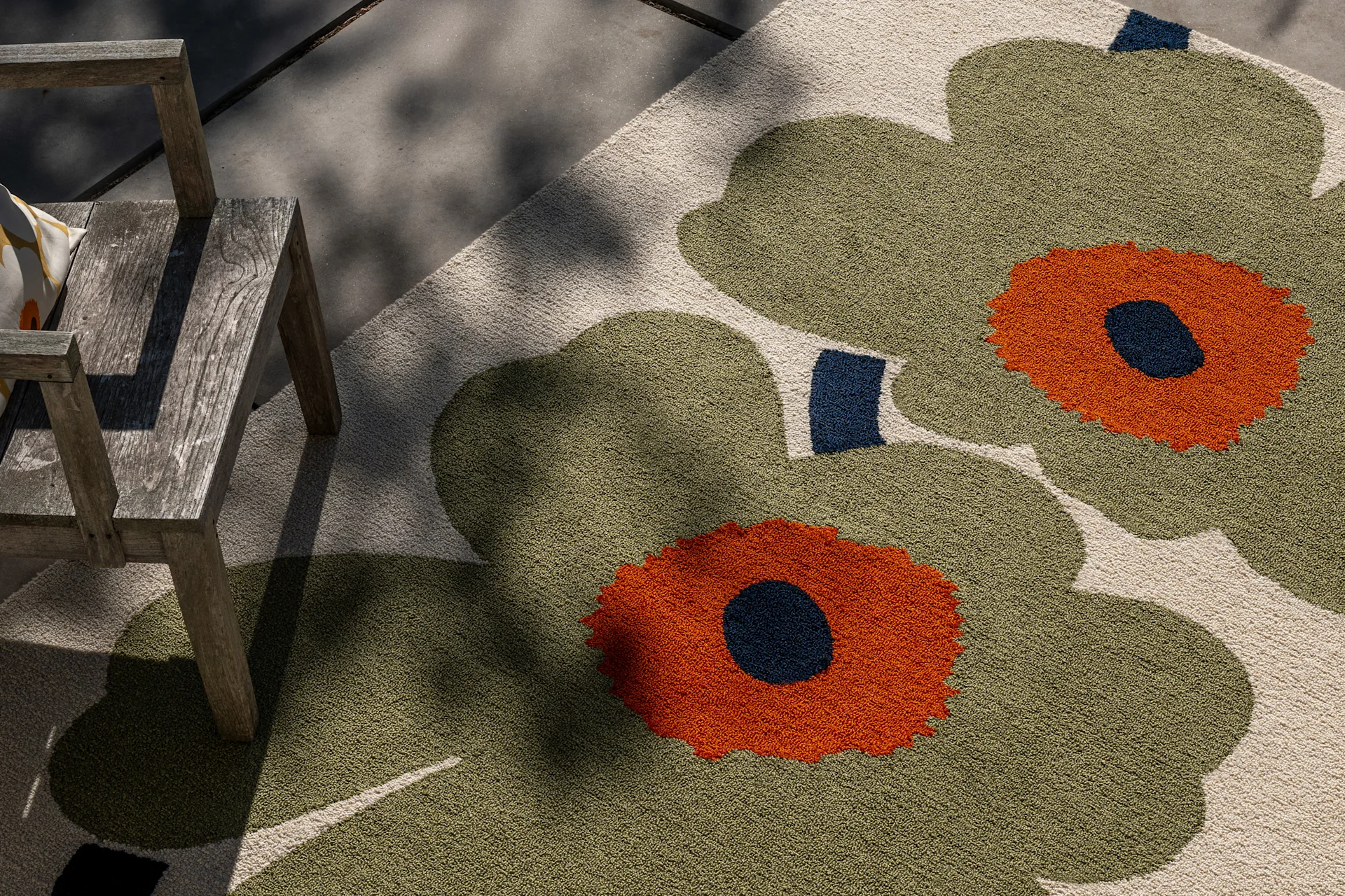 Buitenkleed met groot olijfgroen, oranje en donkerblauw bloemenpatroon, naast een rustieke houten stoel in gevlekt zonlicht.