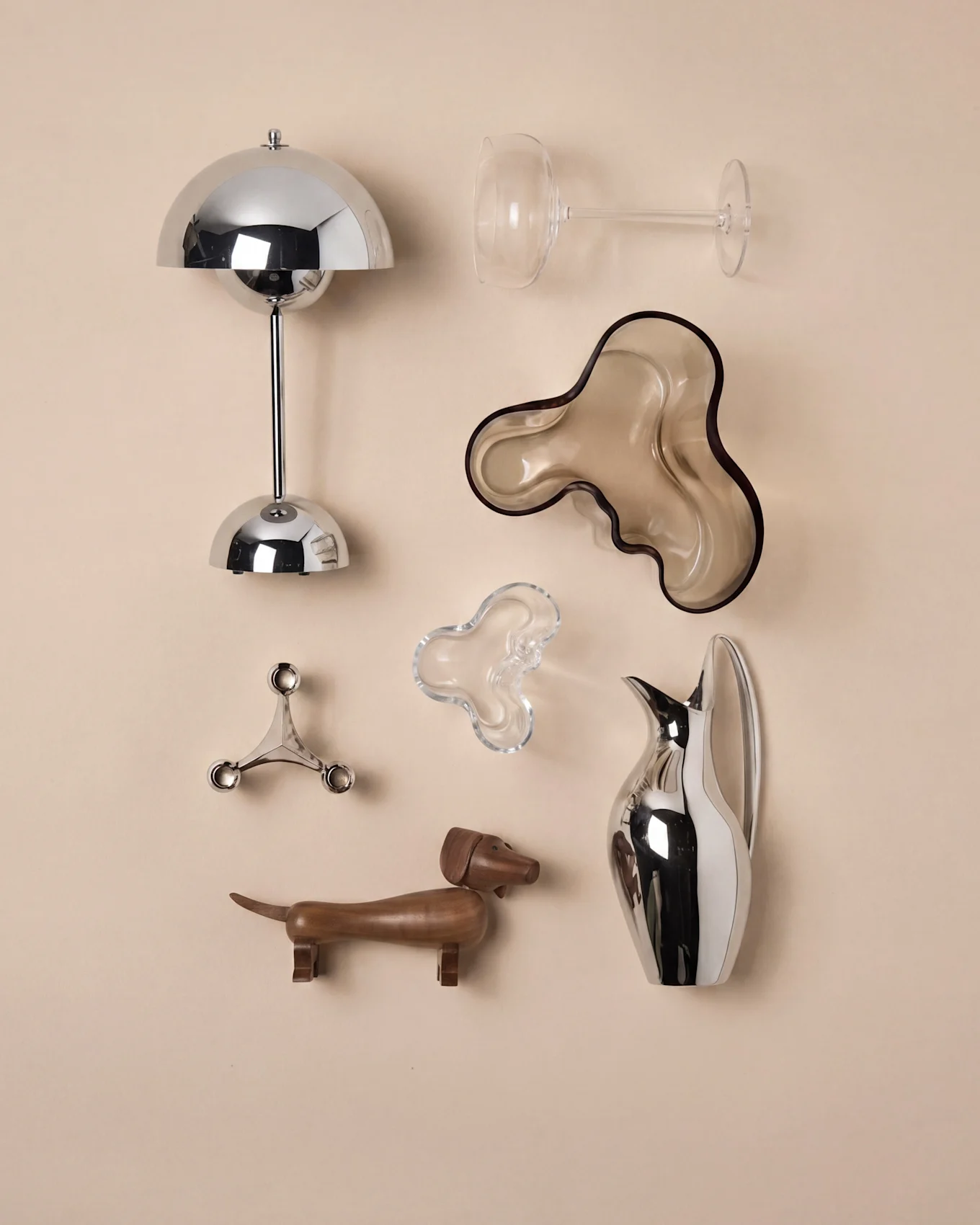 Een arrangement van decoratieve objecten: een chromen lamp, wijnglas, twee organische kommen, een kandelaar, een houten teckel en een kan.