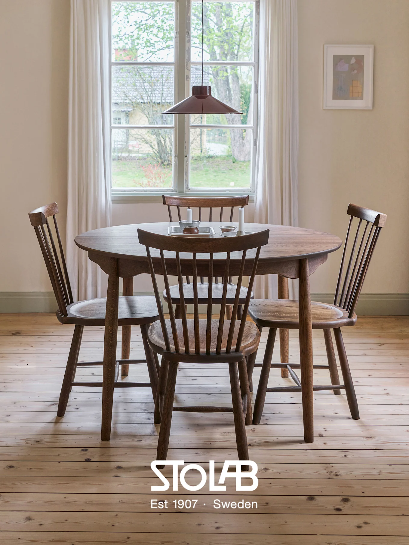 Ronde houten eettafel met vier spijlenstoelen in een lichte kamer, lichte houten vloer, raam en rode hanglamp.