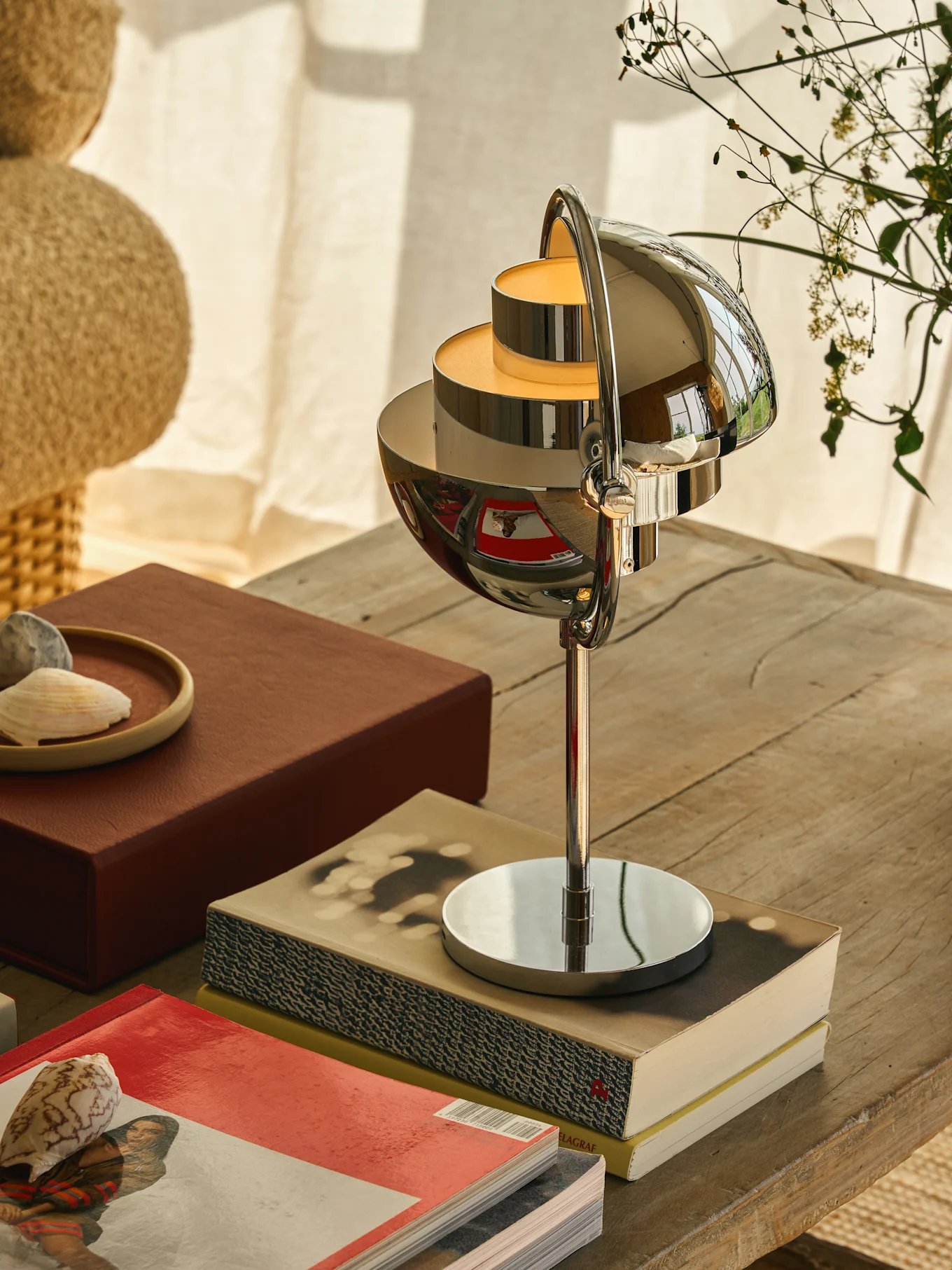 Verchroomde tafellamp met meerlaagse, bolvormige kappen die een warme gloed werpen, op een houten tafel met boeken en schelpen.