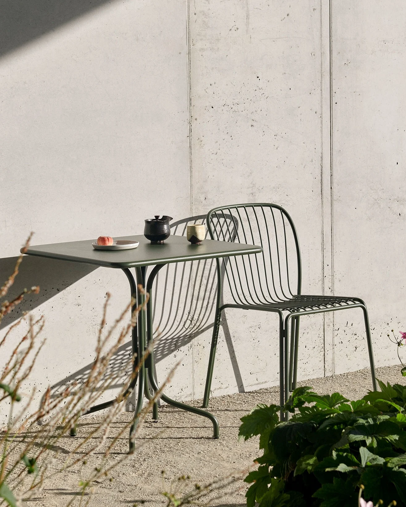 Een donkergroene buitentafel met een perzik, theepot en kop, en een bijpassende lattenstoel, tegen een betonnen muur met sterke schaduwen.
