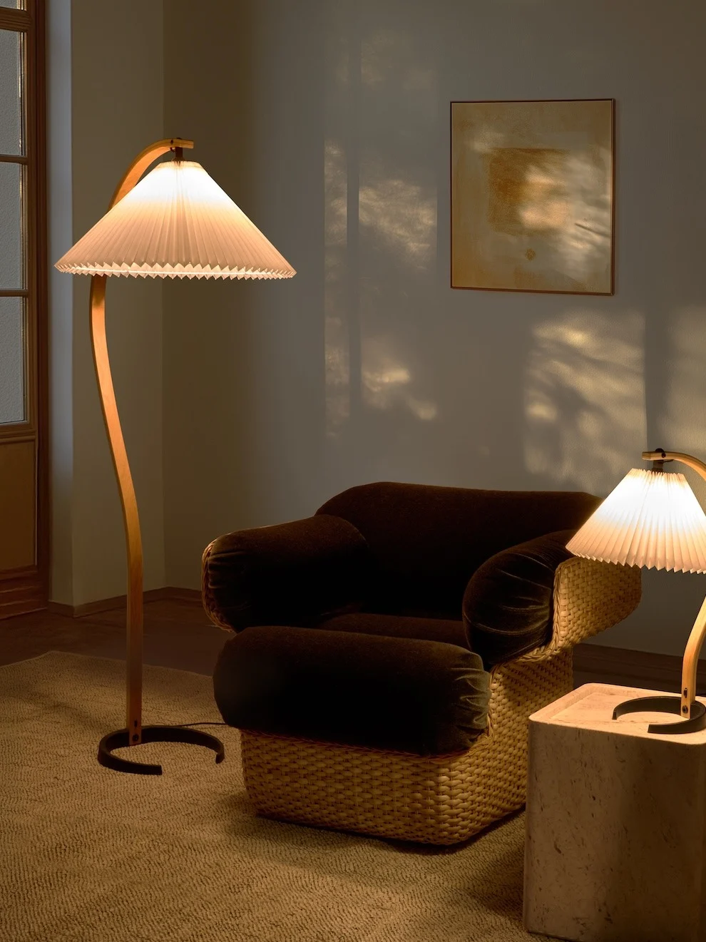 Gezellige kamer met een gebogen vloerlamp, donkere fauteuil met gevlochten onderstel en een kleinere tafellamp. Warm licht creëert schaduwen.
