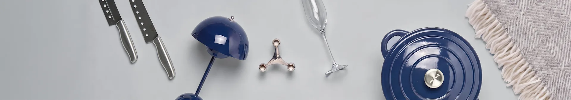 Twee messen, een blauwe lamp, een zilveren houder, een champagneglas, een blauwe pan en een grijs patroon deken op een lichte ondergrond.