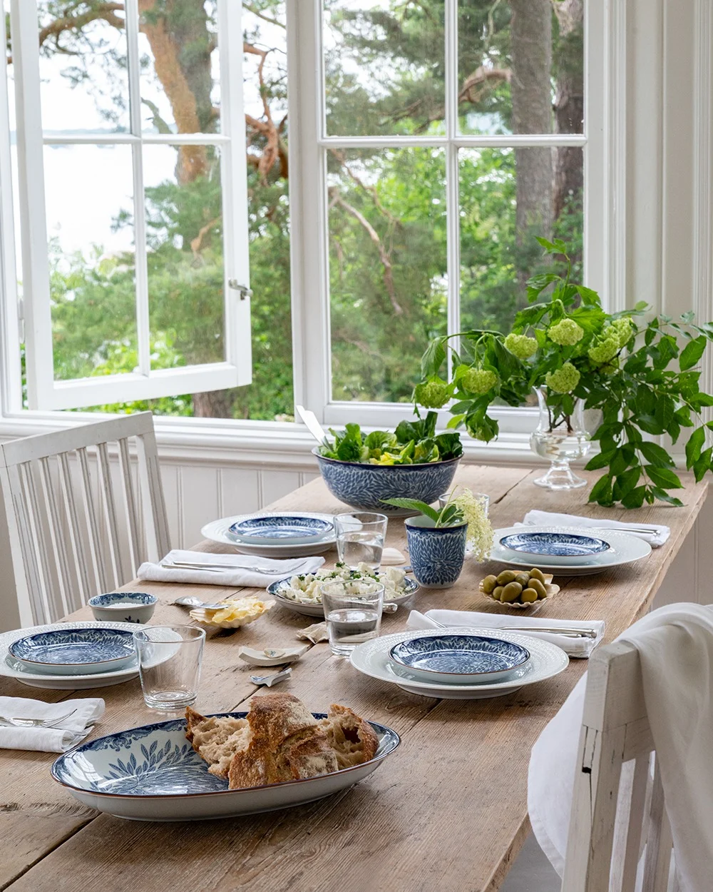 Een rustieke houten eettafel gedekt met blauw-wit servies, salade, brood en olijven, bij een open raam met uitzicht op groene bomen en water.