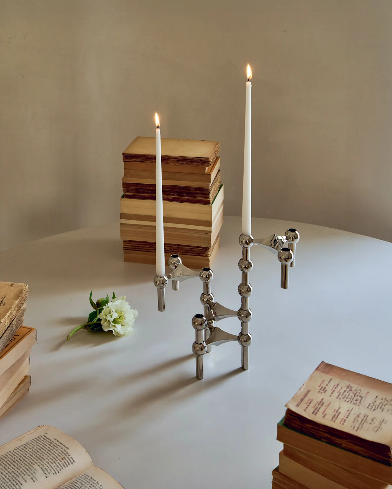 Twee chromen modulaire kandelaars met brandende witte kaarsen, omringd door oude boeken en een witte bloem op een tafel.