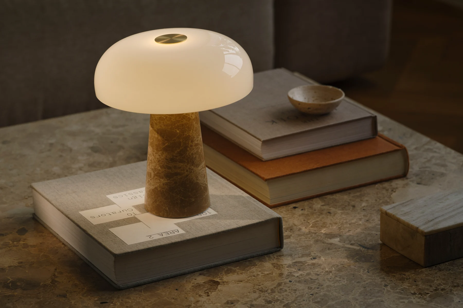 Een paddestoelvormige tafellamp met witte glazen kap en een conische marmeren voet staat op een marmeren tafel met gestapelde boeken.