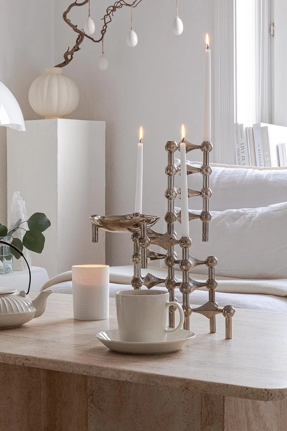 Moderne zilveren kandelaar met drie brandende witte kaarsen, theekopje en theepot op een stenen tafel met paasdecoratie.