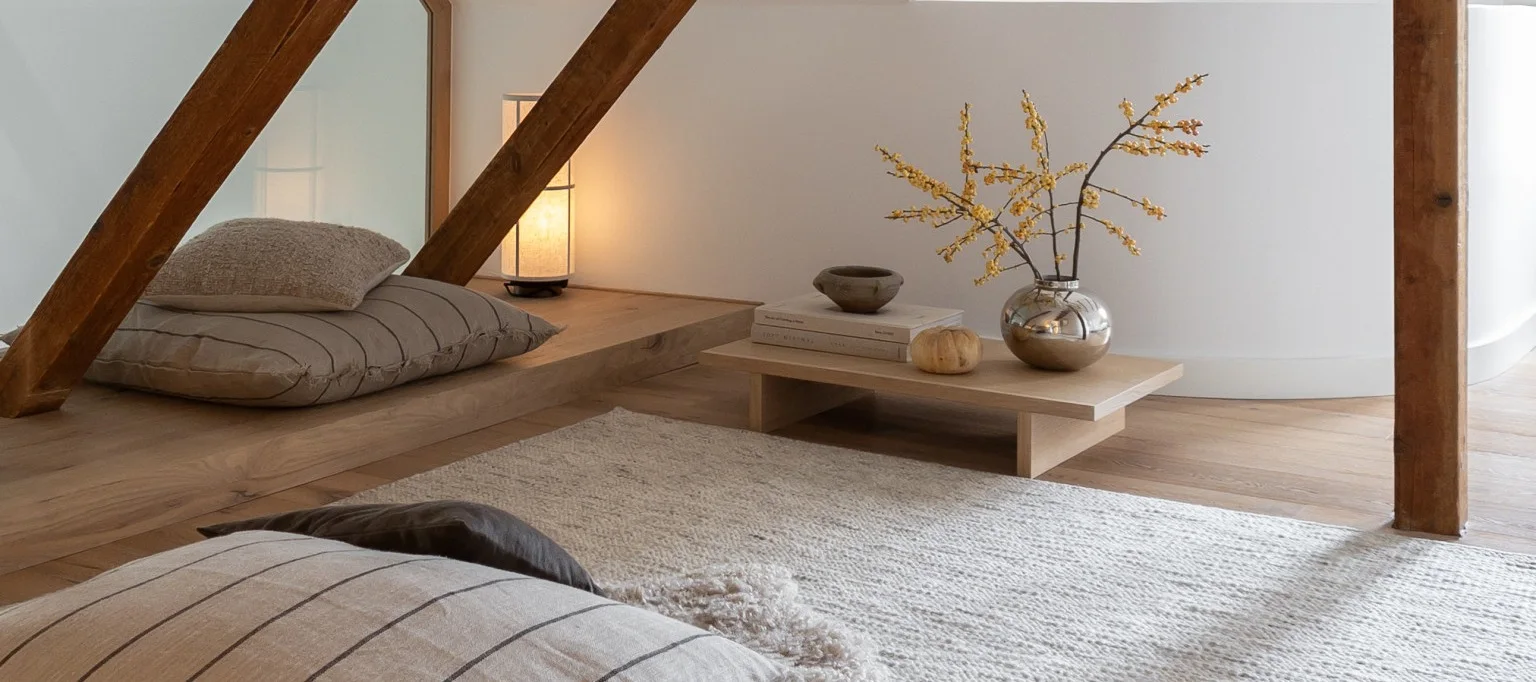 Een gezellige kamer met lichte houten vloeren en witte muren, met vloerkussens, een brandende lamp en een lage tafel met een zilveren vaas met gele bessen.