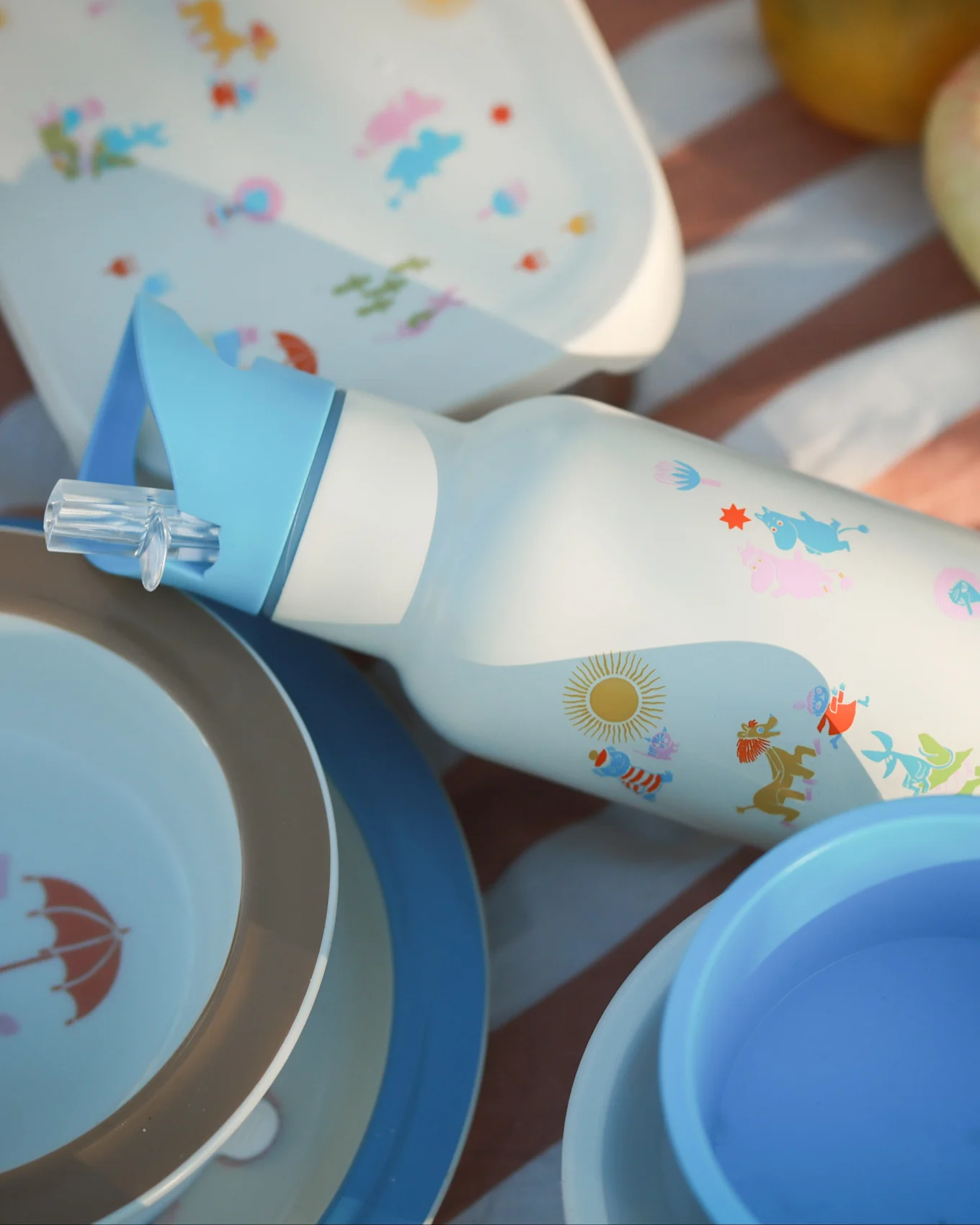 Witte waterfles met blauw rietje, kleurrijke stripfiguren en zon, omringd door bijpassende picknickborden op een gestreepte deken.