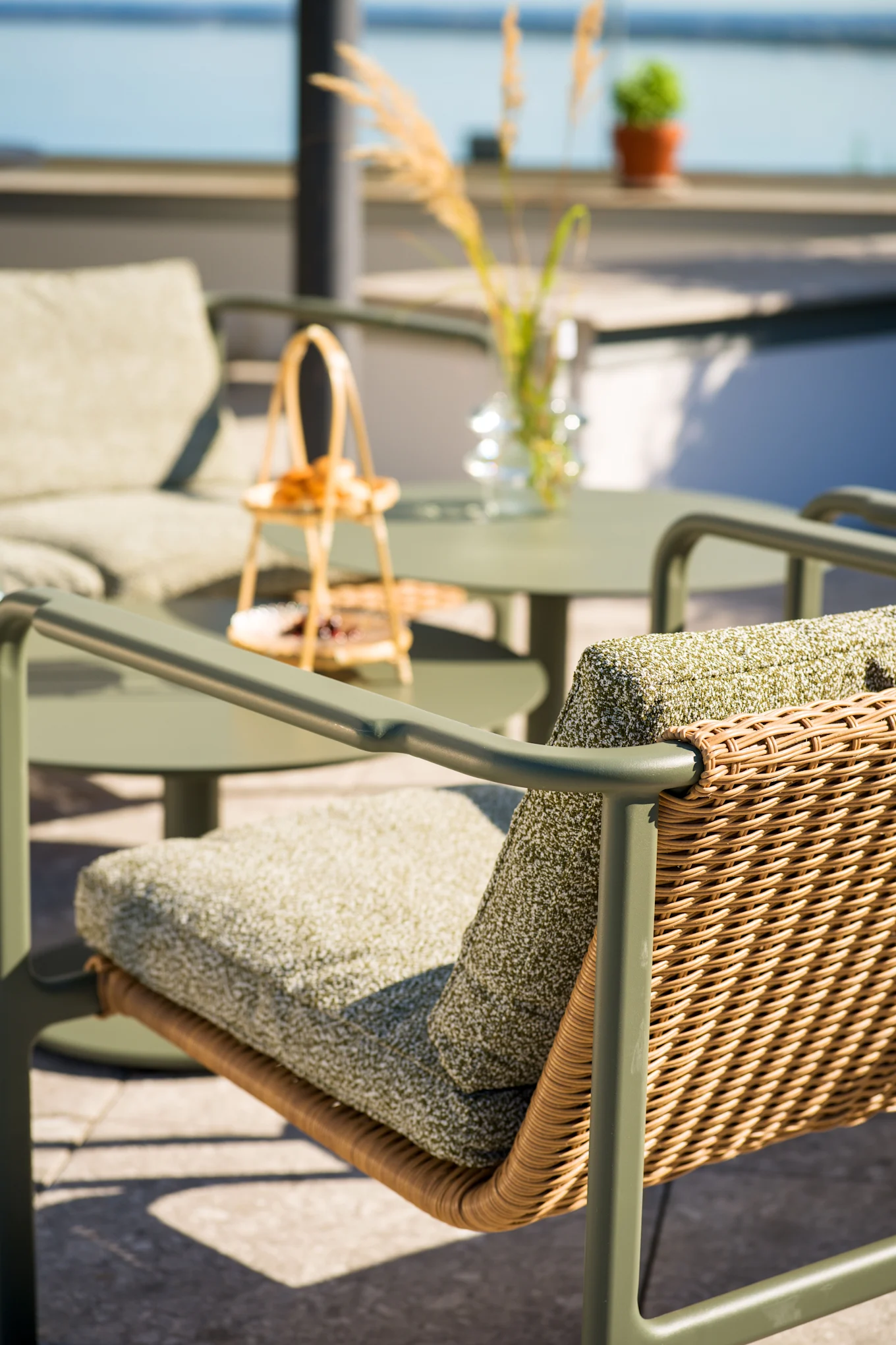 Buitenstoel voor op het terras met olijfgroen frame, gevlochten rotan rugleuning en groene kussens. Bijpassende tafel en stoel op wazige achtergrond bij water.
