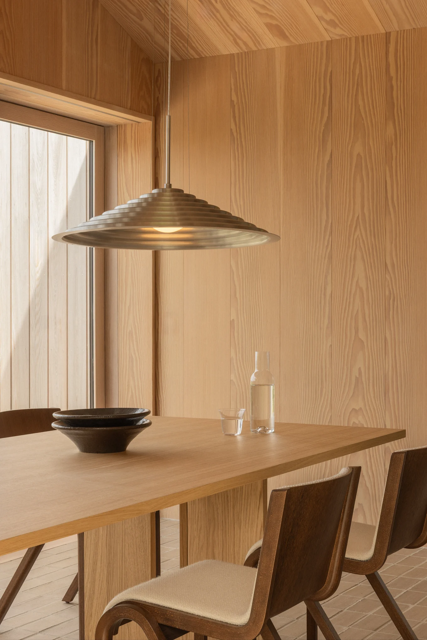 Moderne eetkamer met lichte houten muren, een houten tafel met twee donkere kommen, een waterfles en een gelaagde metalen hanglamp.