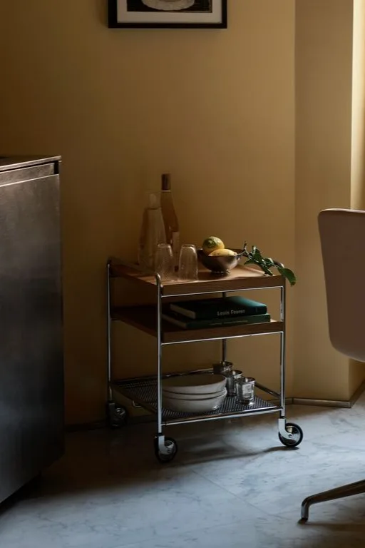 Een drielaags serveerwagen met metalen frame en houten planken, met flessen, glazen, citroenen, boeken en borden.