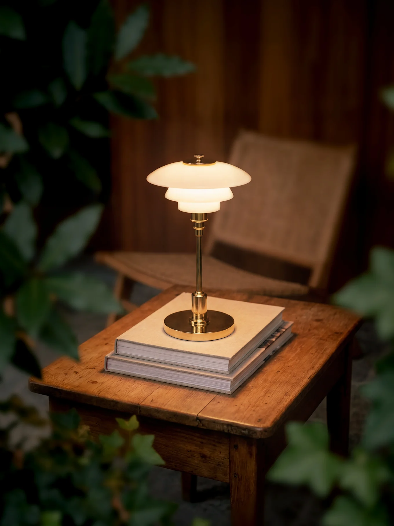 Een gloeiende, meerlaagse messing tafellamp staat op twee boeken op een houten tafel, omringd door donker gebladerte.