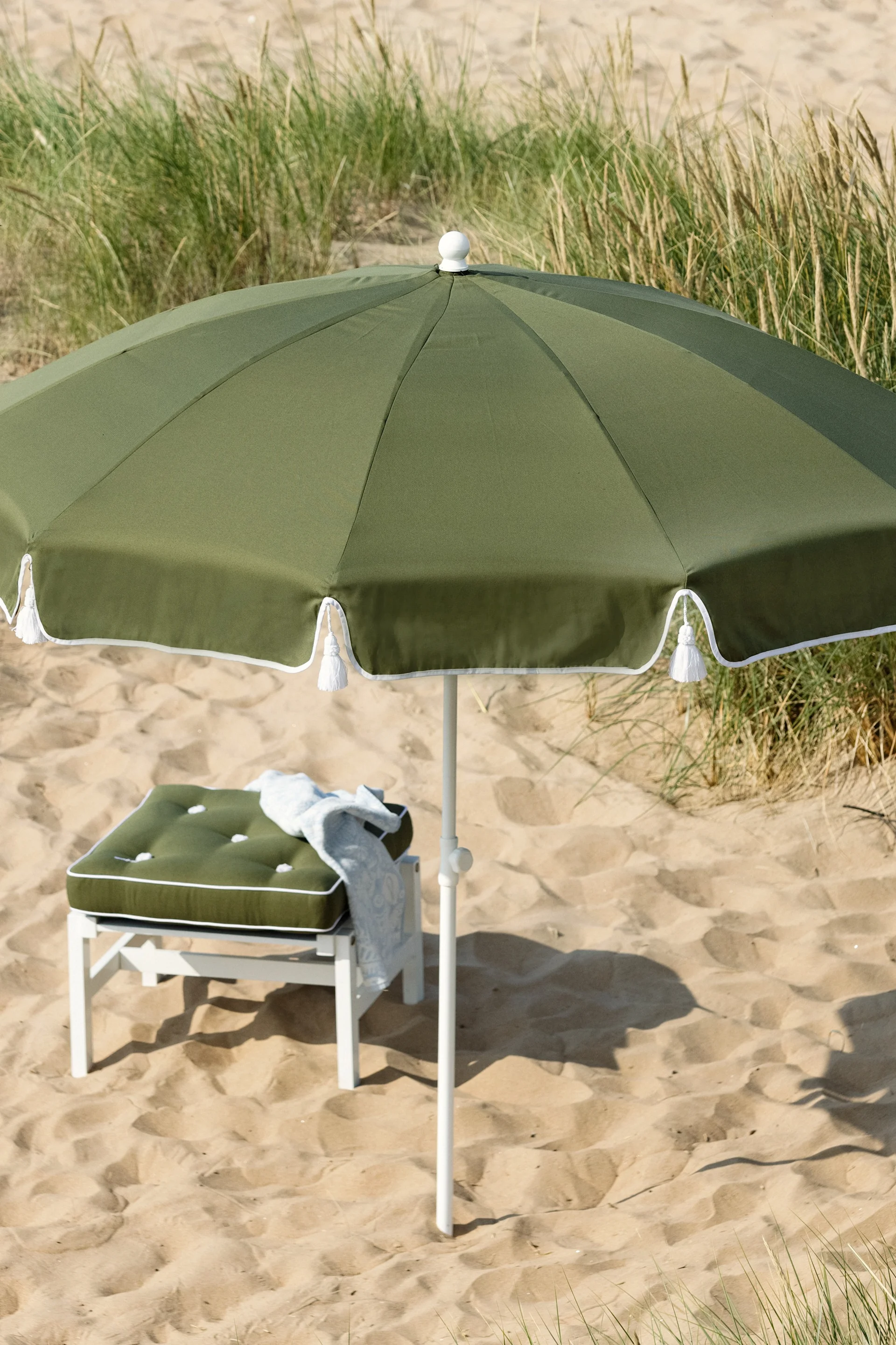 Olijfgroene strandparasol en bijpassend kussen op een witte kruk op een zandstrand met groen gras.