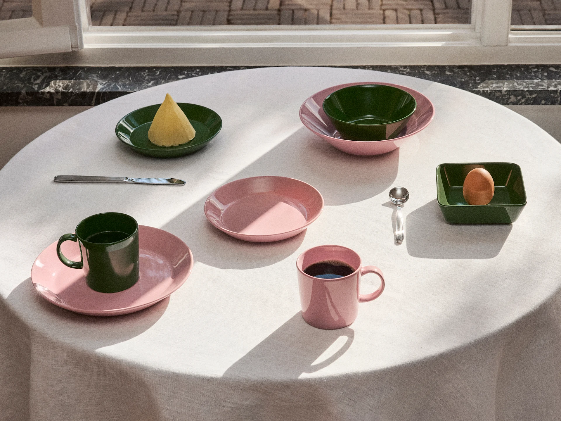Tafel gedekt met roze en donkergroen servies, borden, kommen en mokken, met boter, een ei en koffie onder natuurlijk licht.
