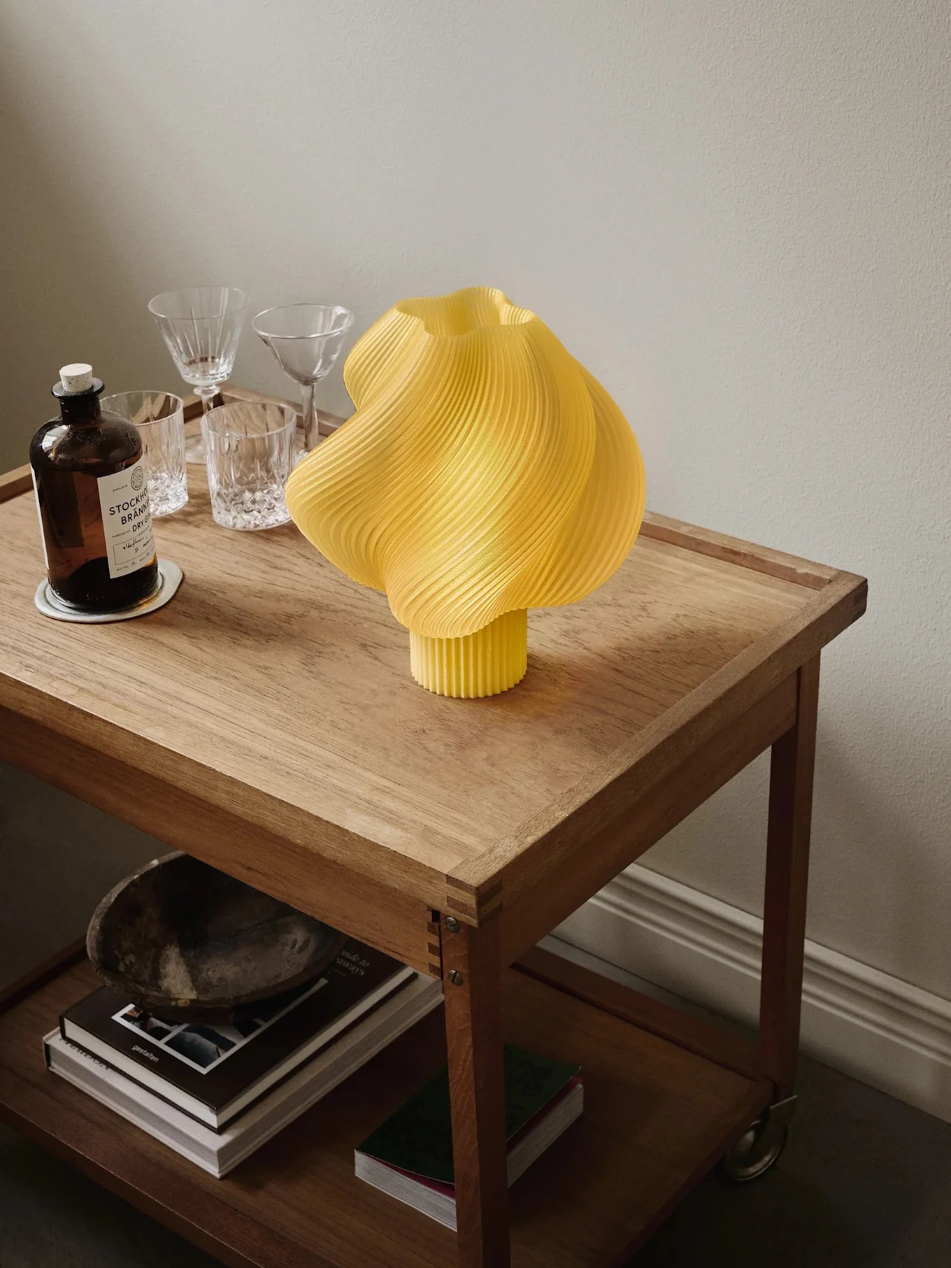 Gele geplooide lamp op een houten trolley met een gin fles, glazen en boeken op de onderste plank.