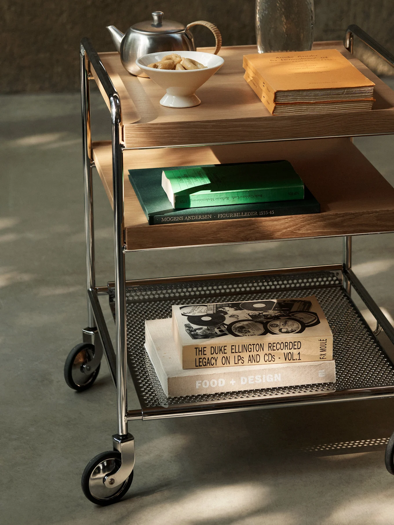 Een drielaags serveerwagen met chromen frame, houten en geperforeerde metalen planken, met boeken, theepot en koekjes.