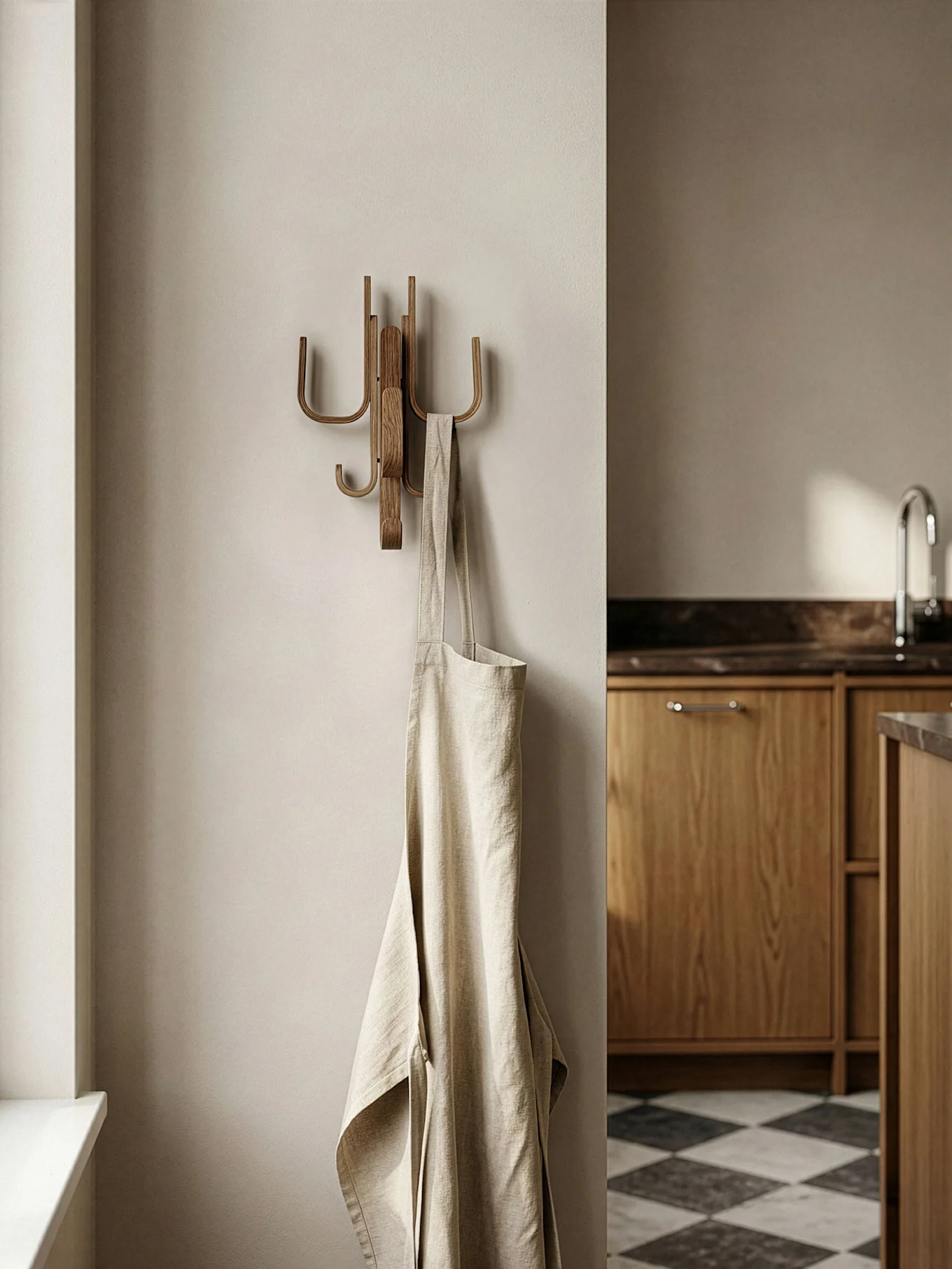 Een lichtbeige linnen schort hangt aan een houten wandhaak in een moderne keuken met houten kasten en een geblokte vloer.