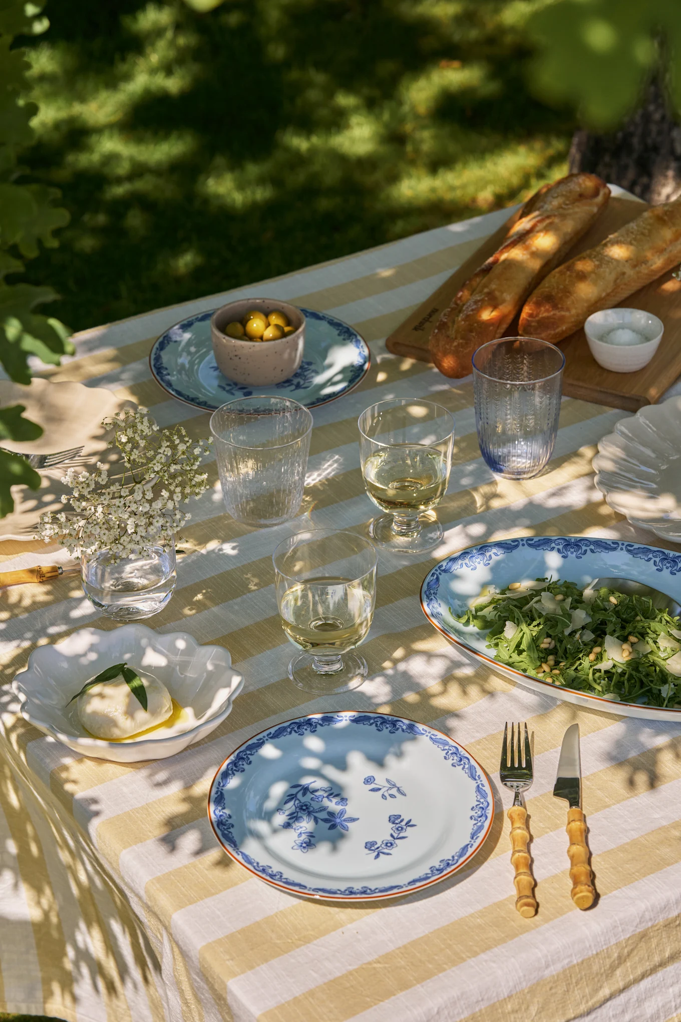 Buiten eettafel gedekt met wit-geel gestreepte tafelkleed, blauwe bloemenborden, wijn, salade, mozzarella, olijven en baguettes.