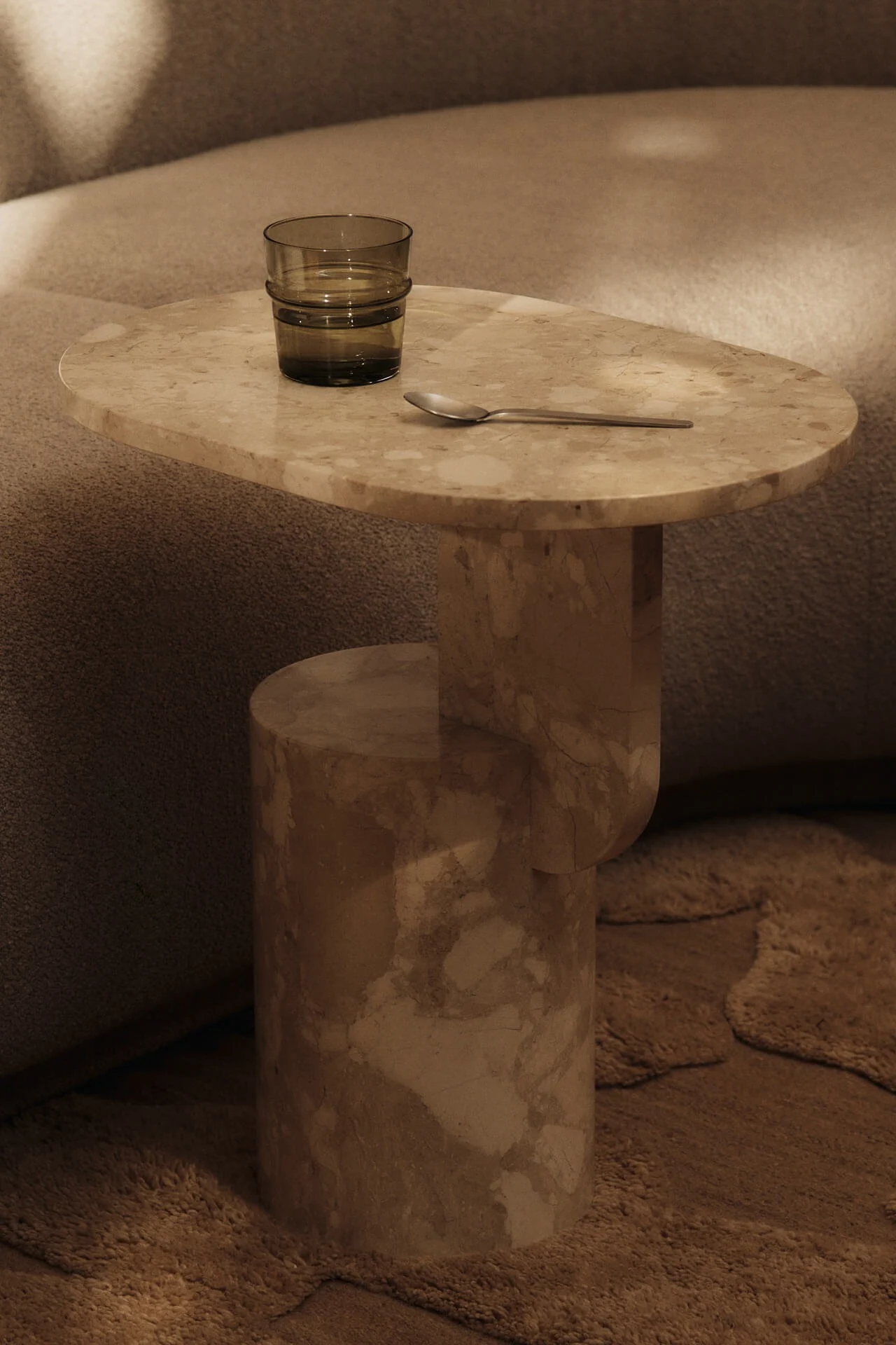 Beige marmeren bijzettafel met een donkergroen glas water en een zilveren lepel, naast een gestructureerde bank en tapijt.