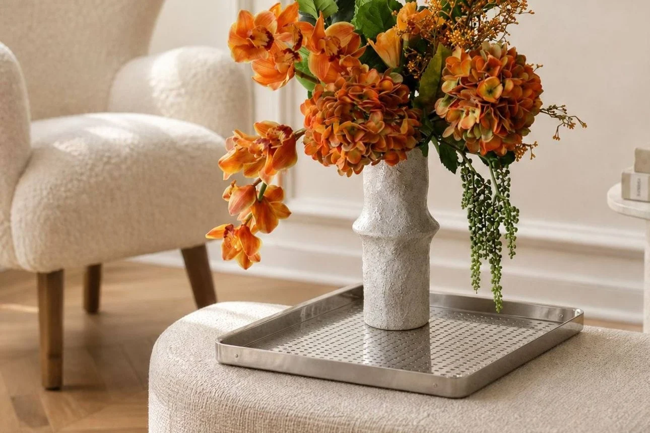 Levendige oranje bloemen in een getextureerde witte vaas op een zilveren dienblad, geplaatst op een lichte poef in een moderne woonkamer.