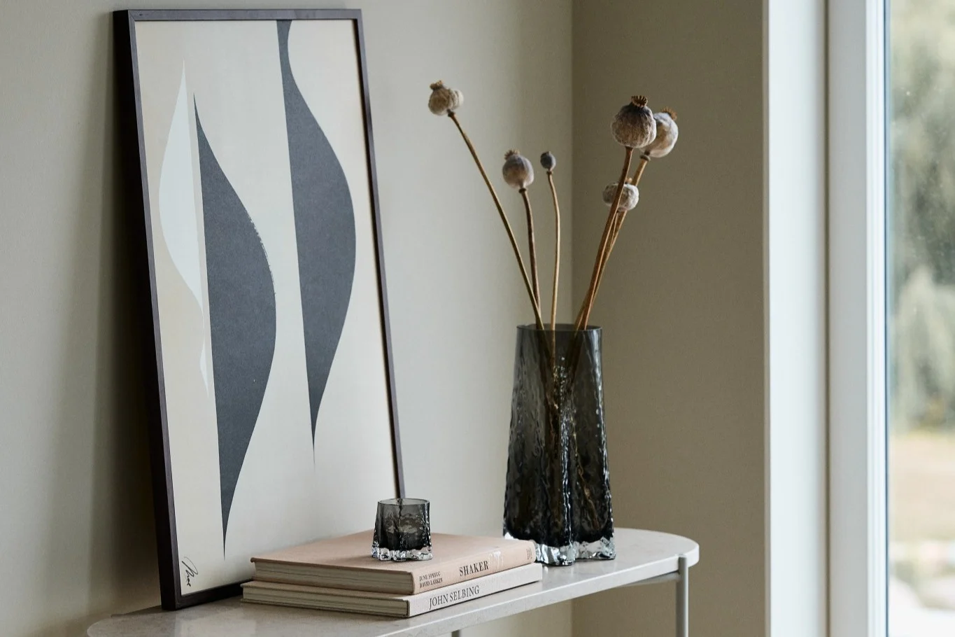 Hier zie je de Gry vaas 30 cm en een Gry kaarsenhouder, beide in de kleur Smoke van Cooee Design, samen met een poster op een bijzettafel. 