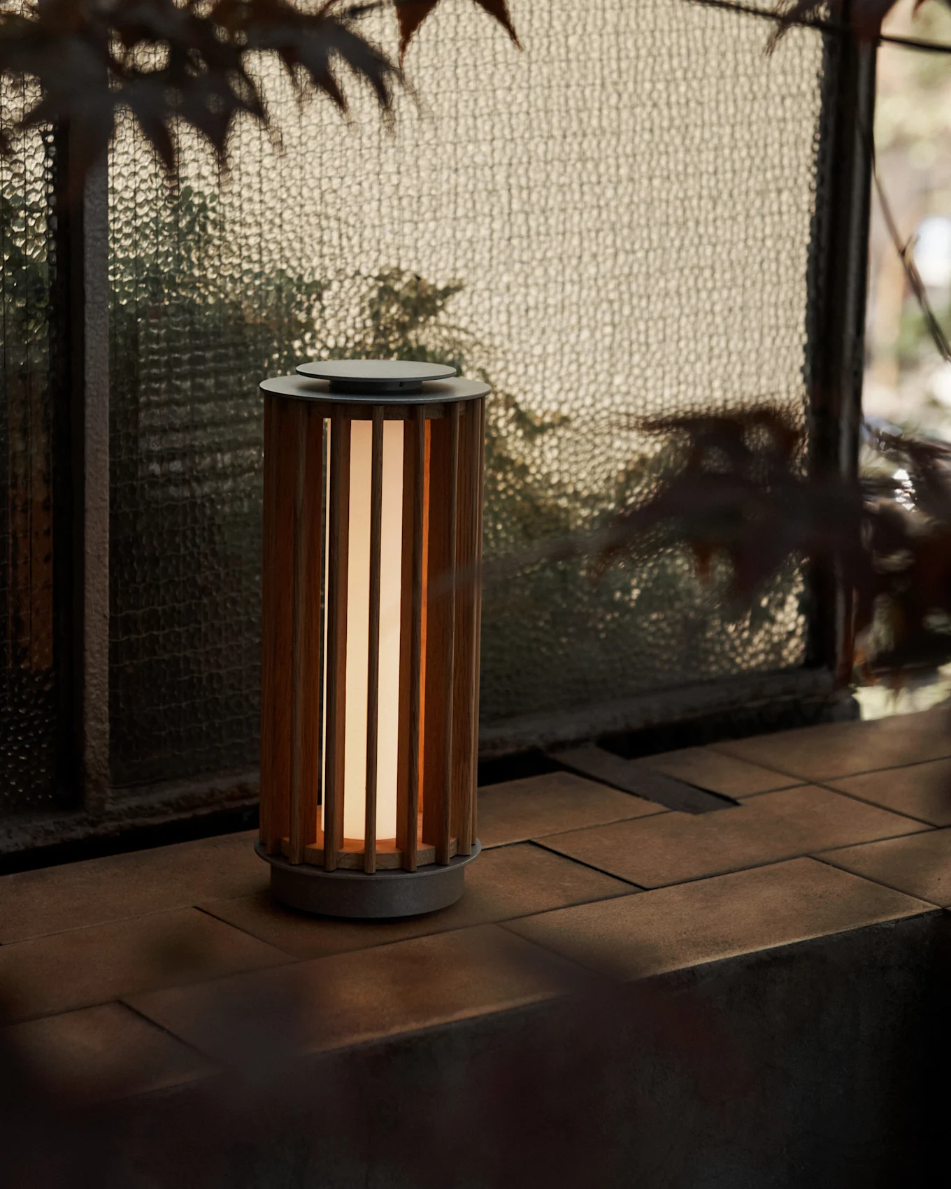 Een cilindervormige draagbare lamp met een houten lamellenbehuizing en warme gloed, op een betegeld oppervlak bij een getextureerd glasraam.