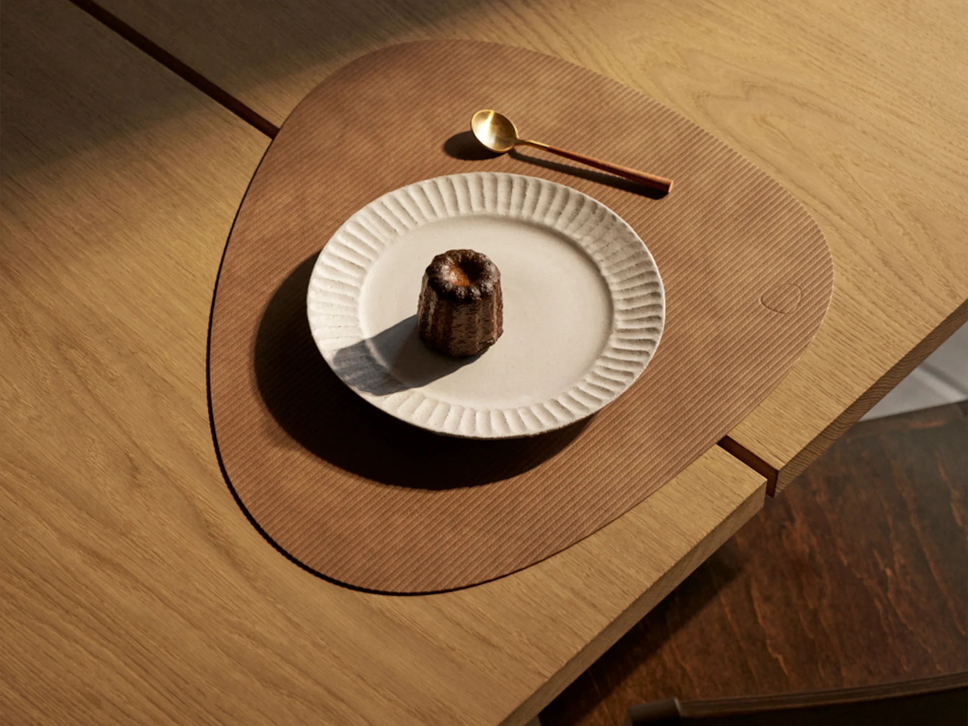 Een canelé-gebakje op een geribbeld bord, een goudkleurige lepel en een driehoekige placemat op een houten tafel, verlicht door zonlicht.