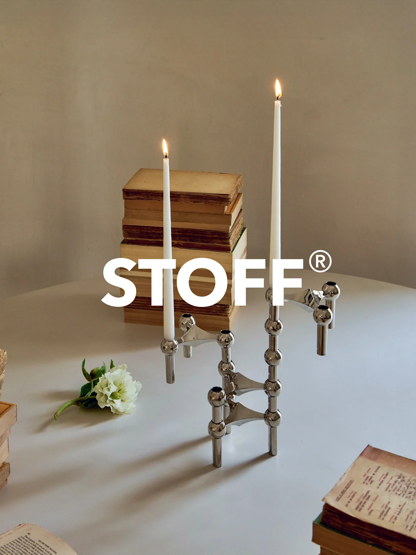 Glanzend chromen modulair kandelaber met twee brandende kaarsen, omringd door oude boeken en een witte bloem op een tafel. STOFF® logo.