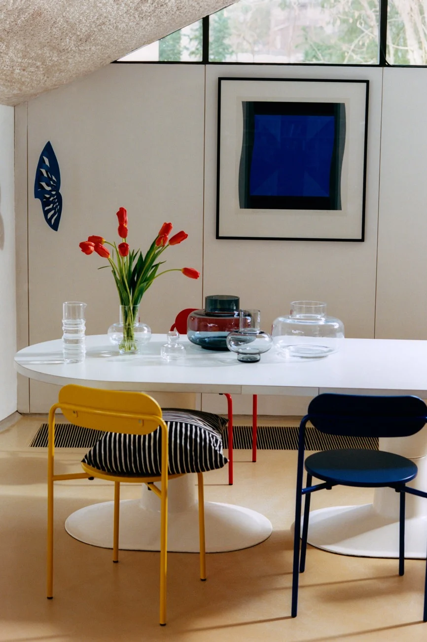 Moderne eetkamer met witte ovale tafel, rode tulpen, glazen vazen, gele en blauwe stoelen en abstracte blauwe kunst.