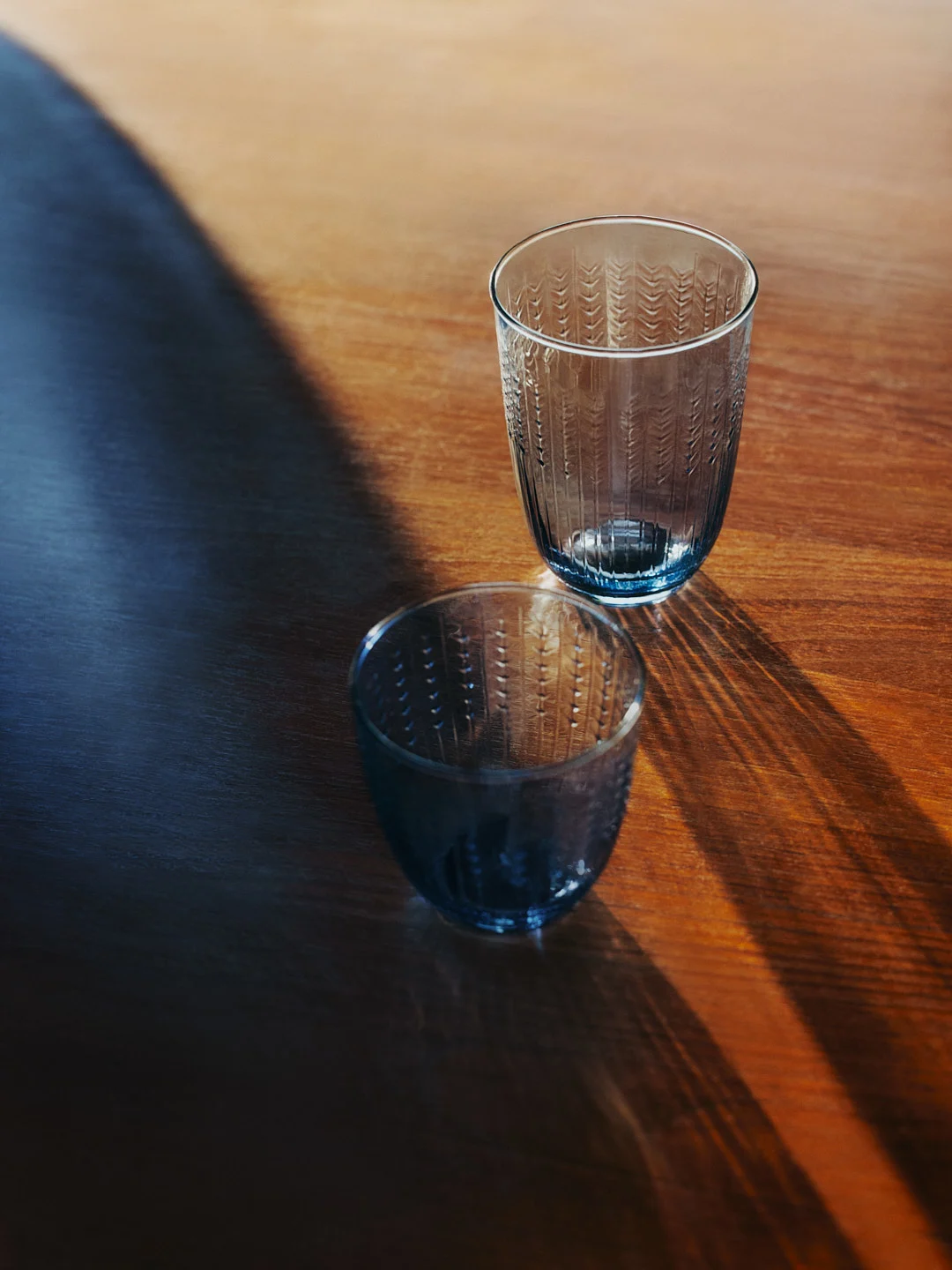 Twee blauwgetinte, gestructureerde glazen op een houten tafel, met lange schaduwen.