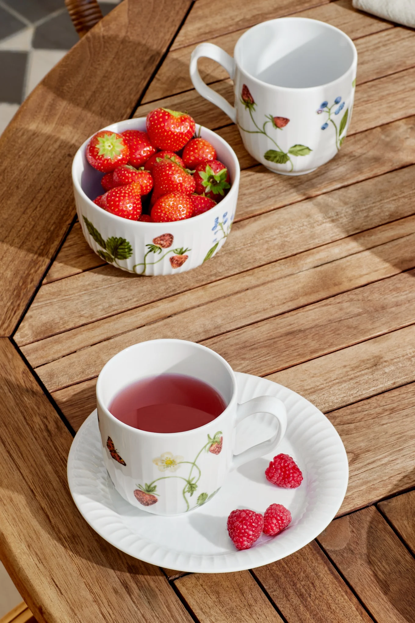 Een witte mok met roze thee en een schotel met frambozen, een kom aardbeien en een lege mok staan op een houten latten tafel in het zonlicht.