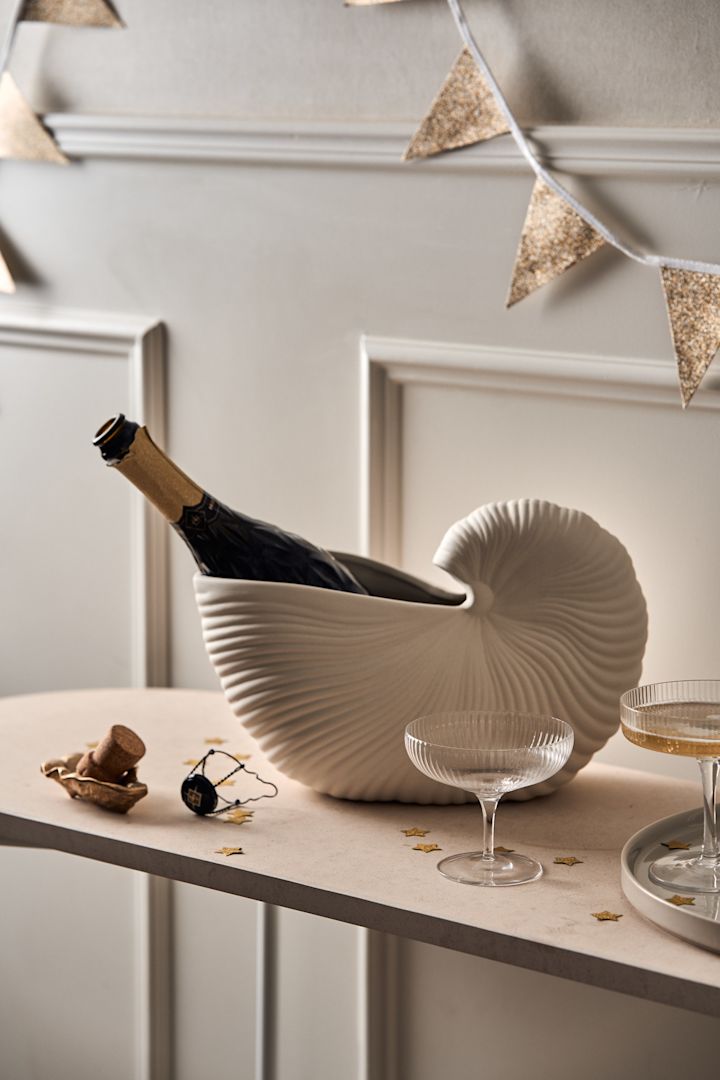 Zo organiseer je een onvergetelijk feest: hier zie je een fles champagne gekoeld in een schelpenvaas van Ferm Living en verschillende champagnecoupes op een tafel als zelfbedieningsdrankstation.