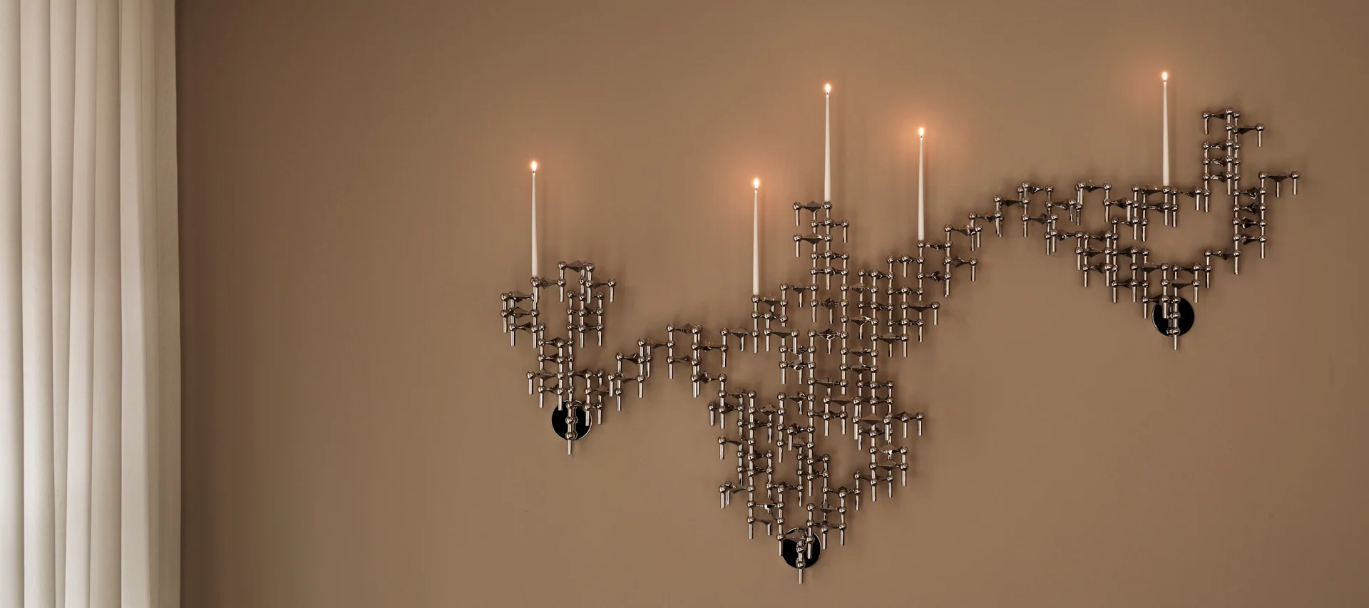 Een wandgemonteerde modulaire zilverkleurige kandelaber met meerdere brandende kaarsen tegen een beige muur, naast verticale jaloezieën.